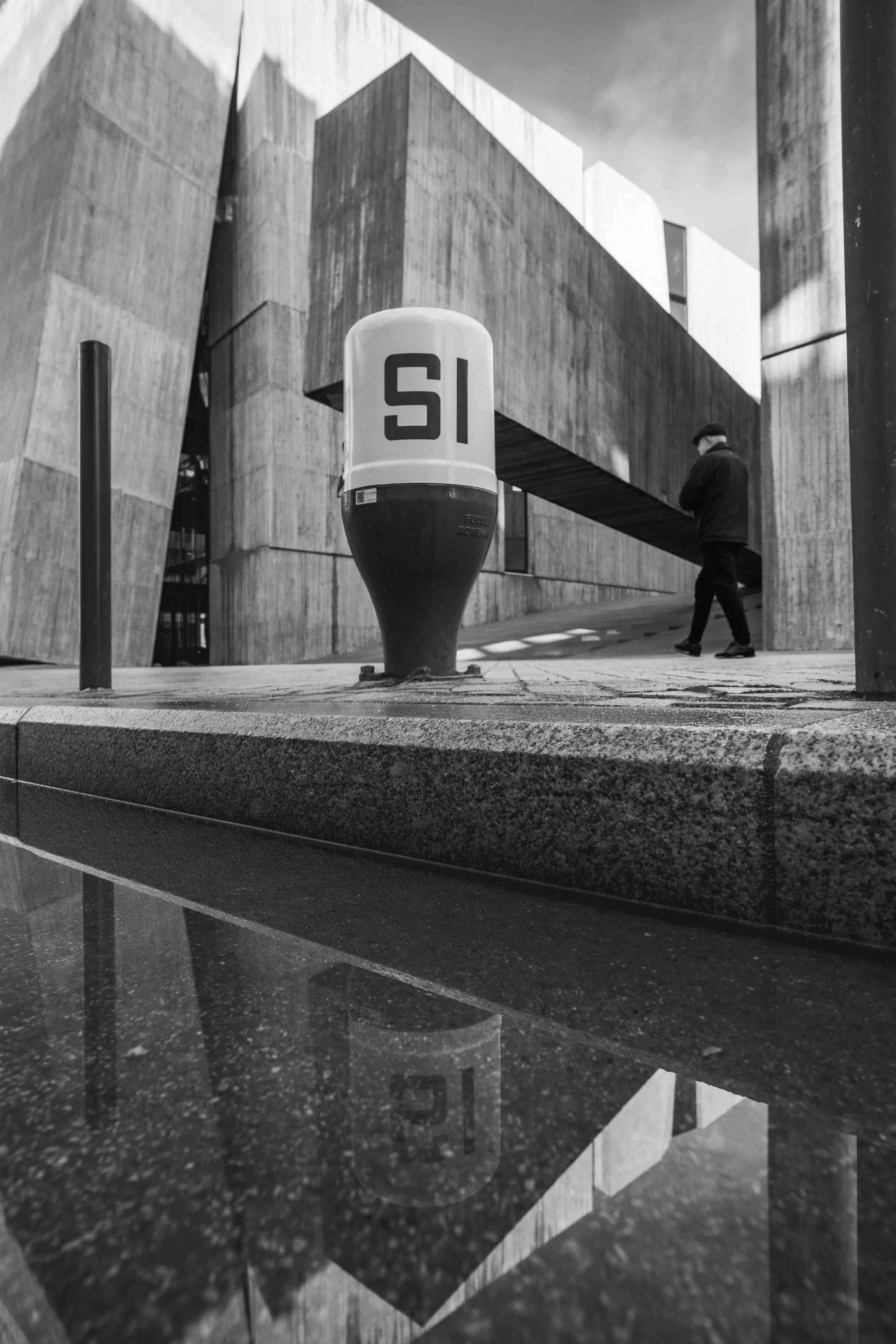 Black and white photo of a modern concrete building with a person walking past. In the foreground, there is a white cylindrical object with the letters 'SI' on it, possibly a sign or marker. The building features sharp angles and geometric design ele