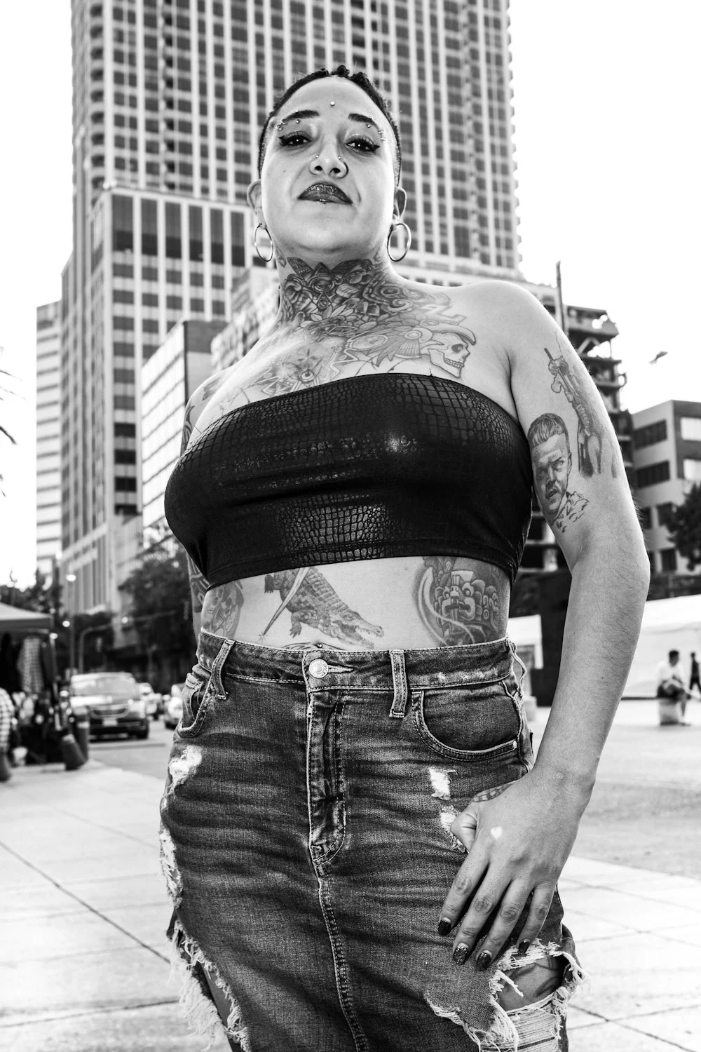 A woman with tattoos standing outdoors in an urban setting with tall buildings behind her, wearing a black strapless top and ripped jeans.