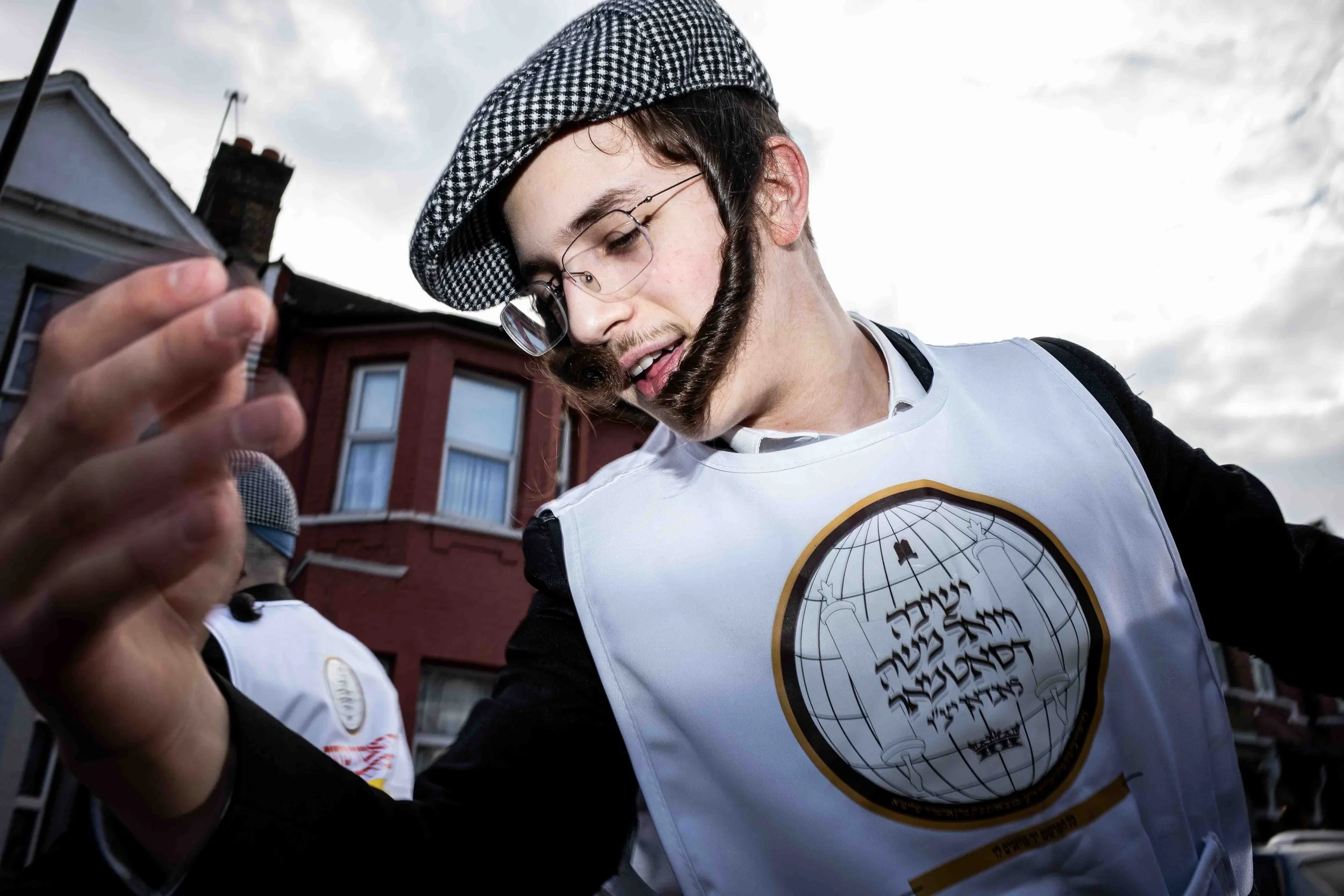A person with curly hair, glasses, and a beard wearing a checkered cap and a white T-shirt with a circular logo, holding a microphone. In the background, there are buildings and cloudy sky.