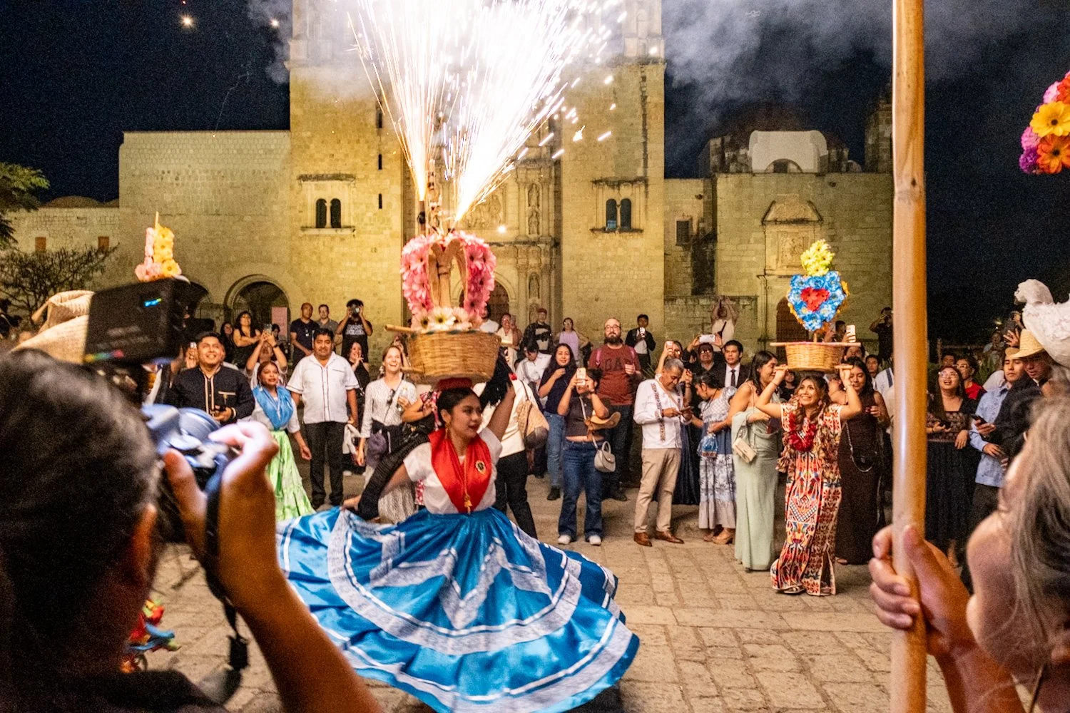 People celebrating at night with traditional dancers, fireworks, and decorated baskets, in front of an historic stone building.