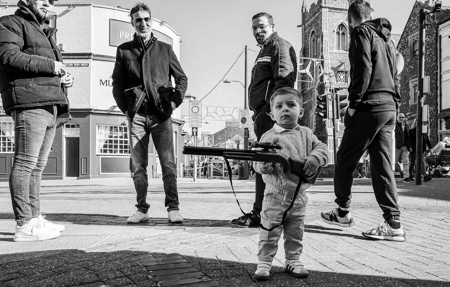 A young boy holding a toy gun standing on a city street, with four adults gathered behind him near a church, on a sunny day.