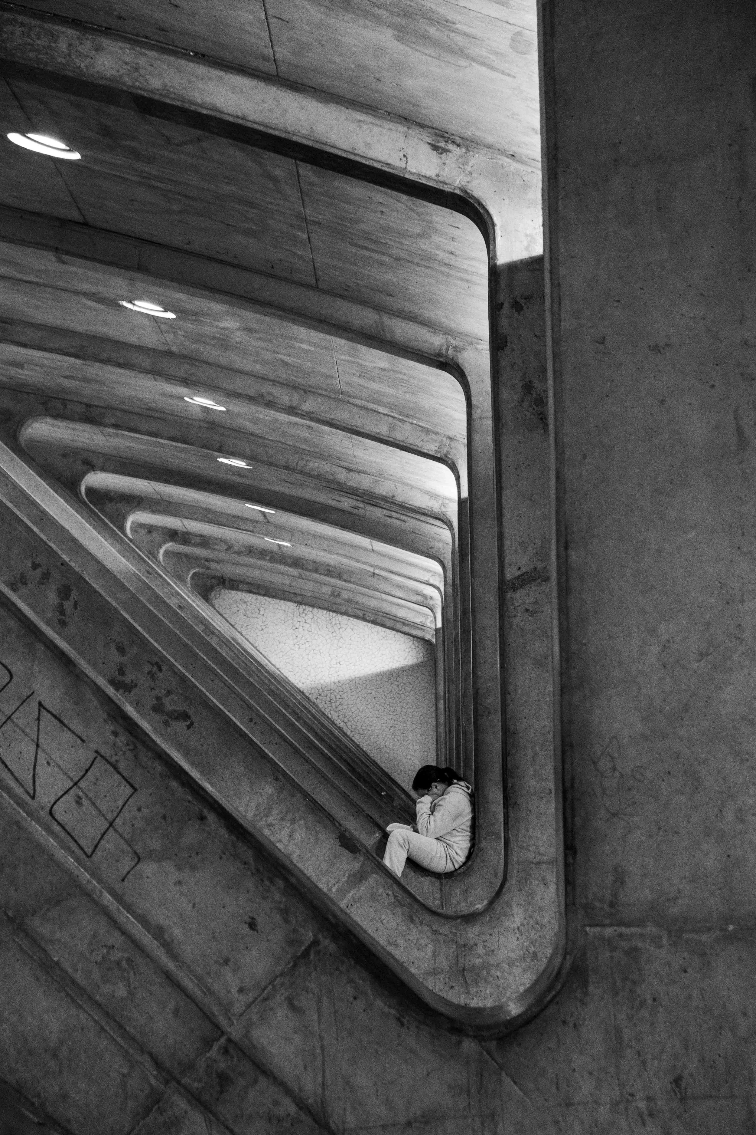 A person sitting on the ledge of a stairwell landing, reading a book or using a device, with repetitive archways of the staircase visible in black and white.