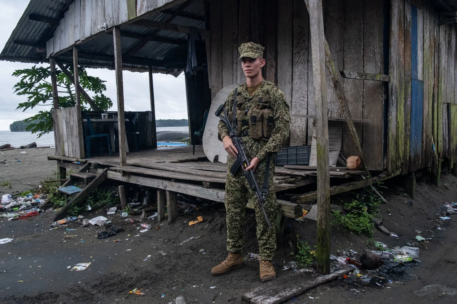 A soldier in camouflage uniform standing on a sandy area in front of a wooden hut, holding a rifle with both hands. The hut appears weathered with some furniture visible inside, and there is scattered trash on the ground.