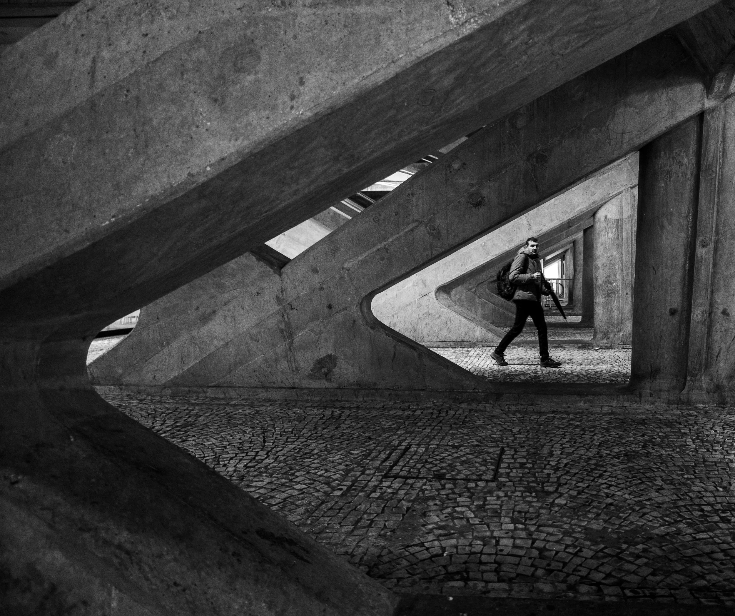 A man with a backpack holding a long object while walking on a cobblestone street under an overpass with large concrete beams overhead.
