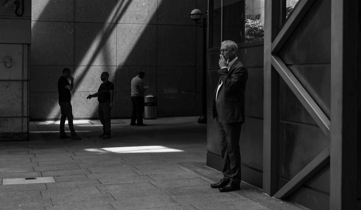 A man in a suit stands against a wall, while three other people stand further away in a cityscape, engaging with each other, with shadows cast on the wall.