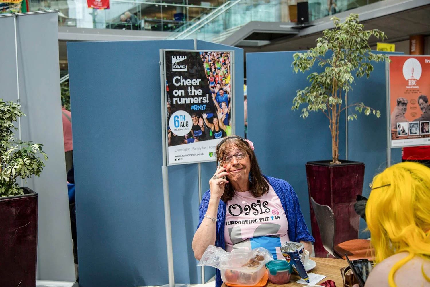 A woman with glasses and long brown hair wearing a pink Oasis t-shirt and a blue cardigan, sitting at a table with food containers and snacks, in an indoor public space with a plant and posters behind her.