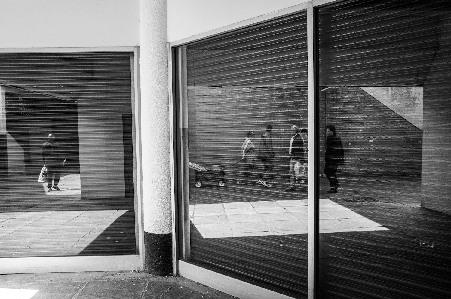 Reflection of people walking outside, visible through glass windows with horizontal blinds, in black and white.