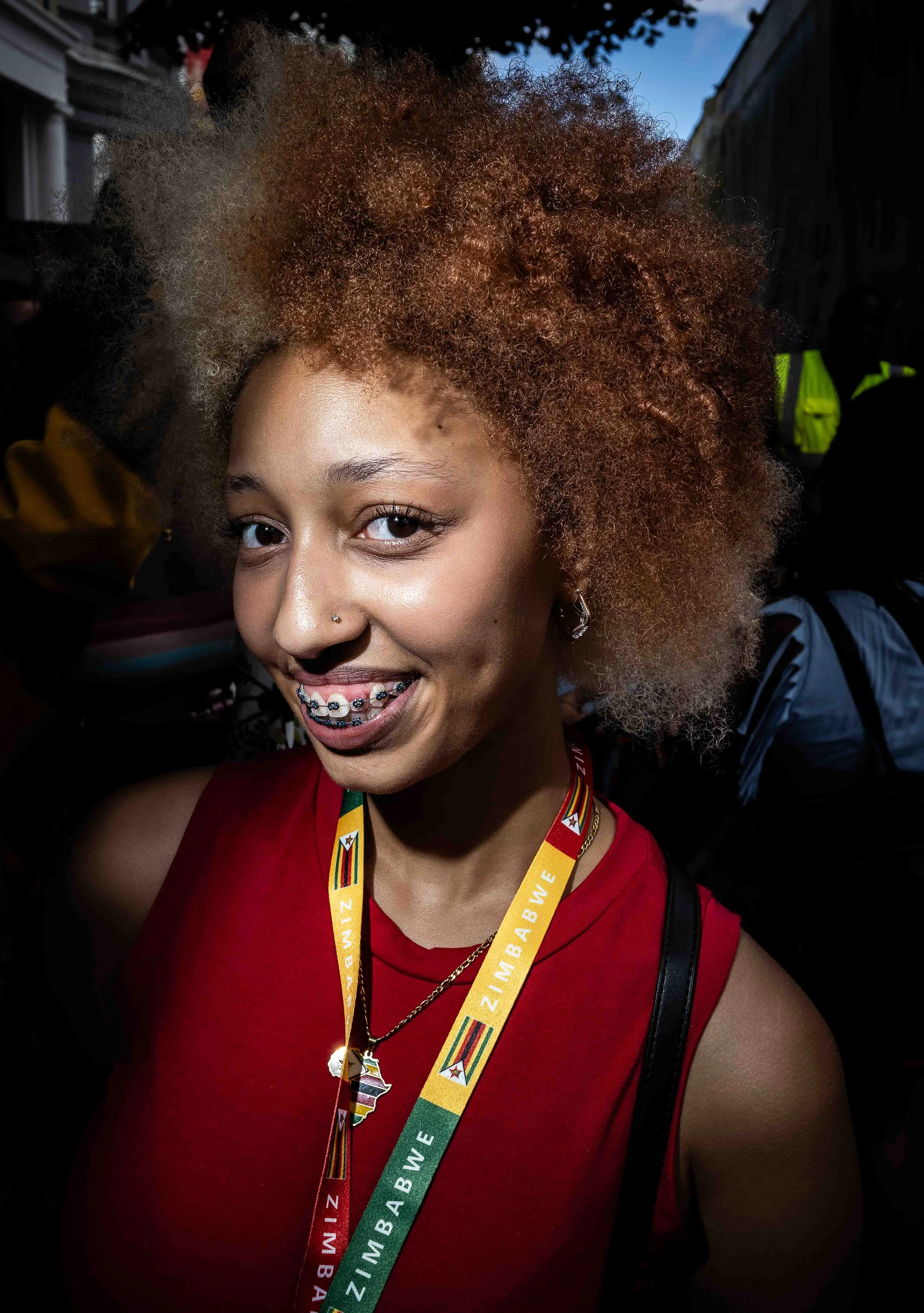 A young woman with light brown skin and curly, two-toned hair, smiling at the camera. She wears a red top, a black earring, a nose piercing, and a medal lanyard with the words 'BIMBA BWB' and flags of Zimbabwe. In the background, there are people and
