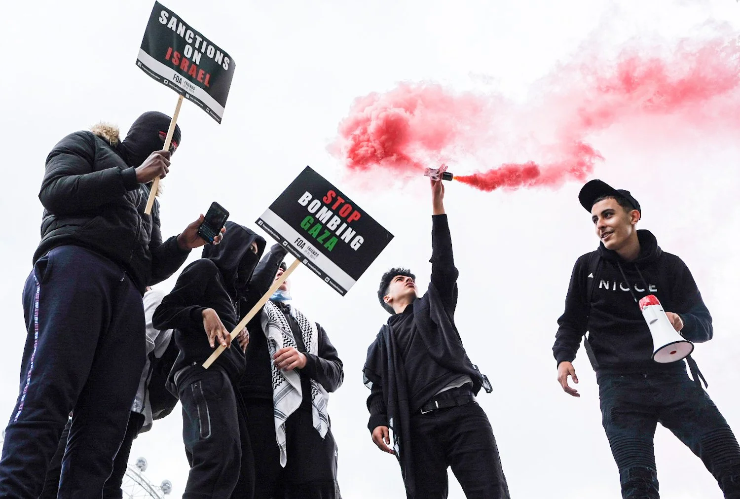 A group of protesters holding signs that say 'Sanctions on Israel' and 'Stop bombing Gaza.' One protester is holding a red smoke flare, and a person is holding a megaphone.