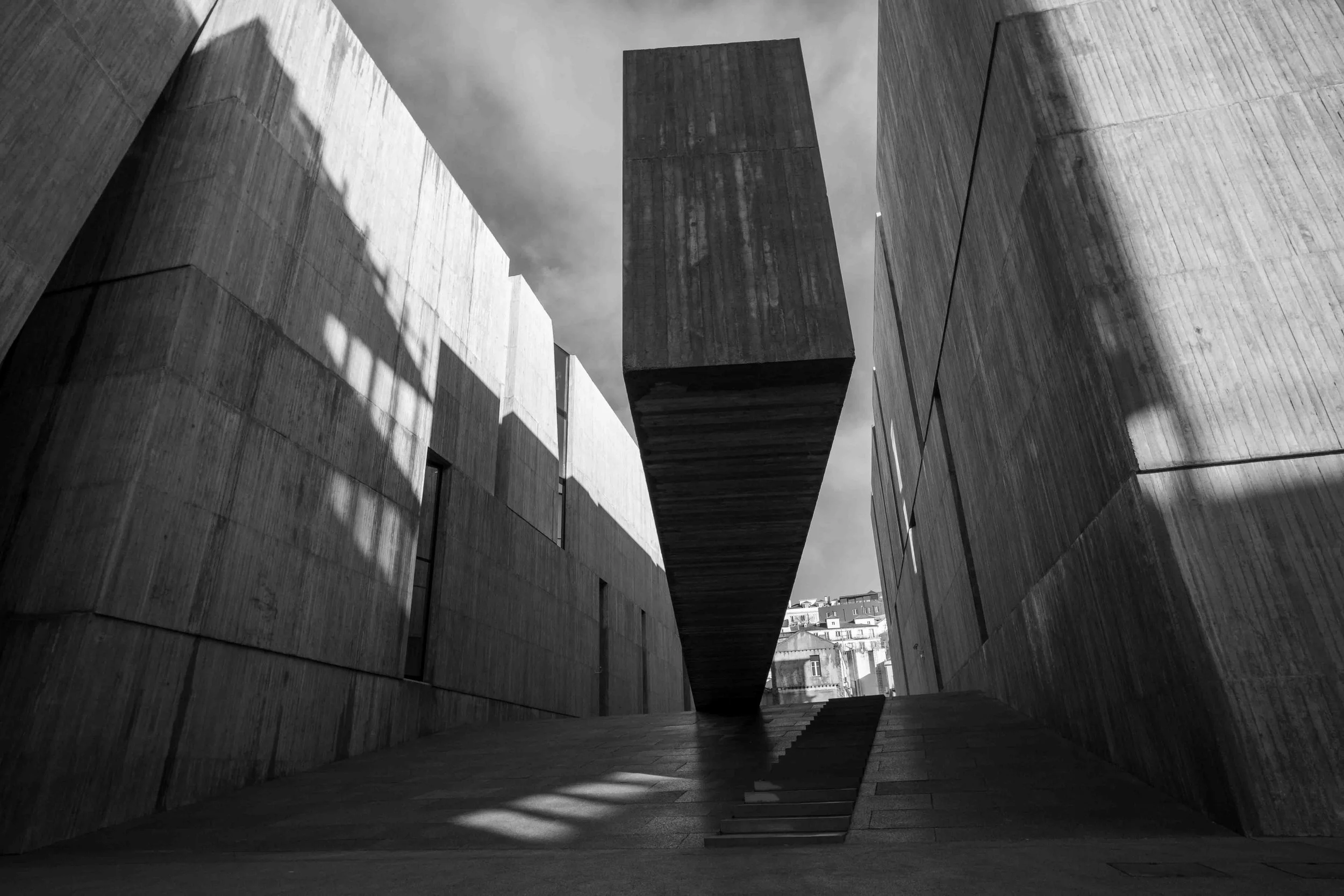 Black and white photo of modern buildings with concrete walls and angular shapes, featuring a large suspended rectangular block in the center, casting shadows on the ground and adjacent structures, with a glimpse of city buildings in the background.