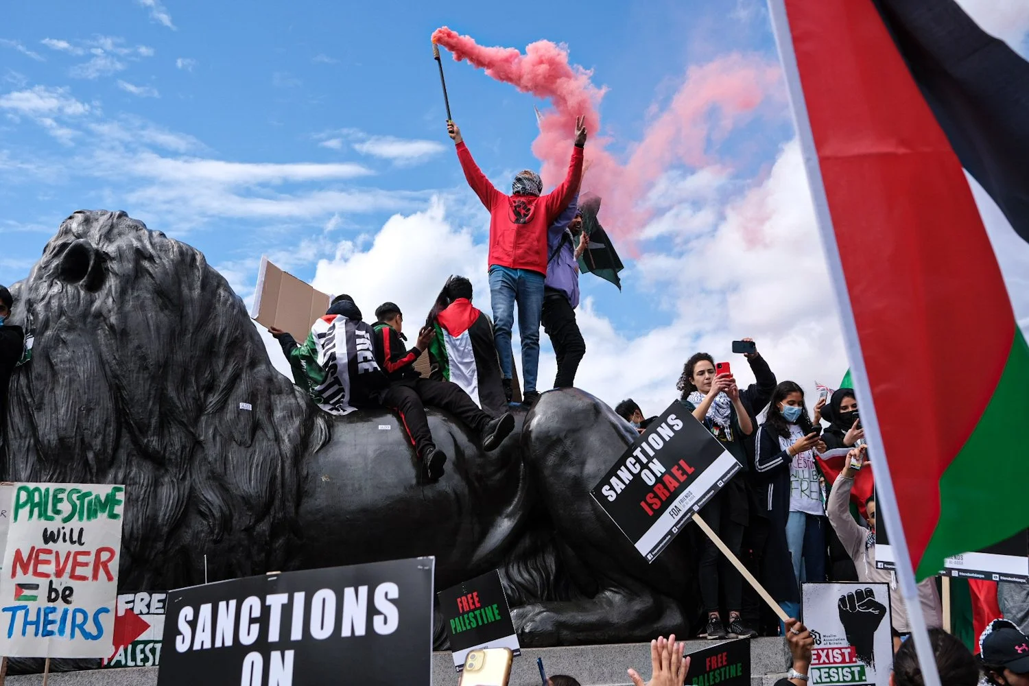 Protesters gather around a large statue of a lion, holding signs and banners supporting Palestine and calling for sanctions on Israel. One person raises a pink smoke flare while others take photos or hold flags, amid a clear sky with scattered clouds