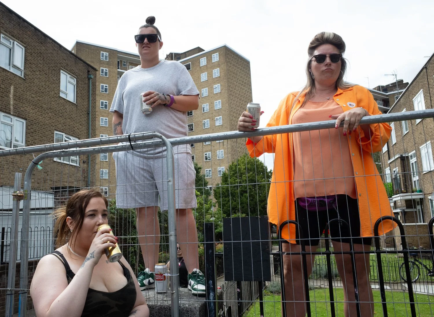 Three women drinking beer outside in an urban area, with an apartment building in the background. One woman is sitting on the ground, another is standing on a fence, and the third is leaning on the fence.