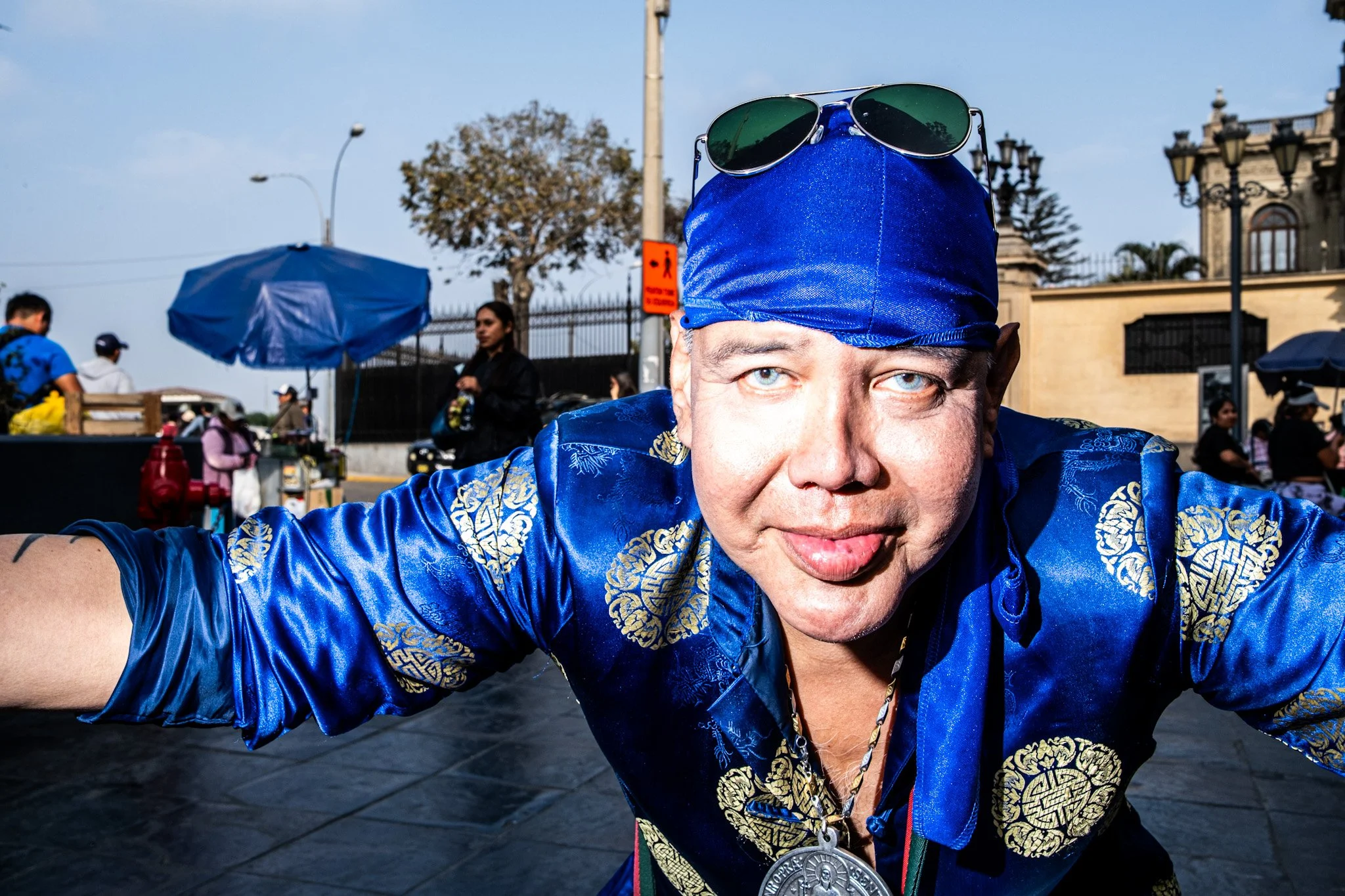 A man wearing a shiny blue traditional outfit with gold patterns and a blue head covering. He has sunglasses on his head and a large silver medal around his neck. The background shows people and street vendors with umbrellas, and a decorated building