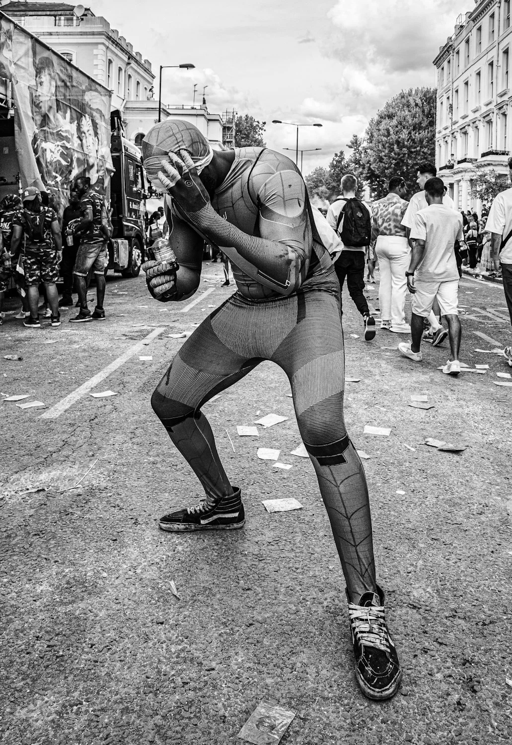 A person in a Spider-Man costume dancing or posing on a city street, with a crowd of people and police officers in the background. The scene appears lively with scattered debris on the ground.
