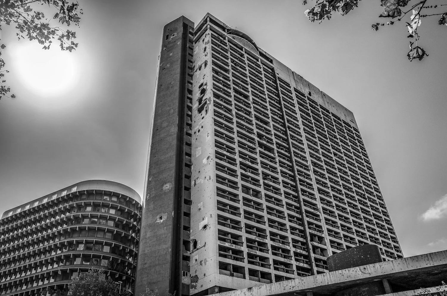 Black and white photo of two tall, abandoned concrete buildings with broken windows and exposed structure, with tree branches and the sun in the sky.