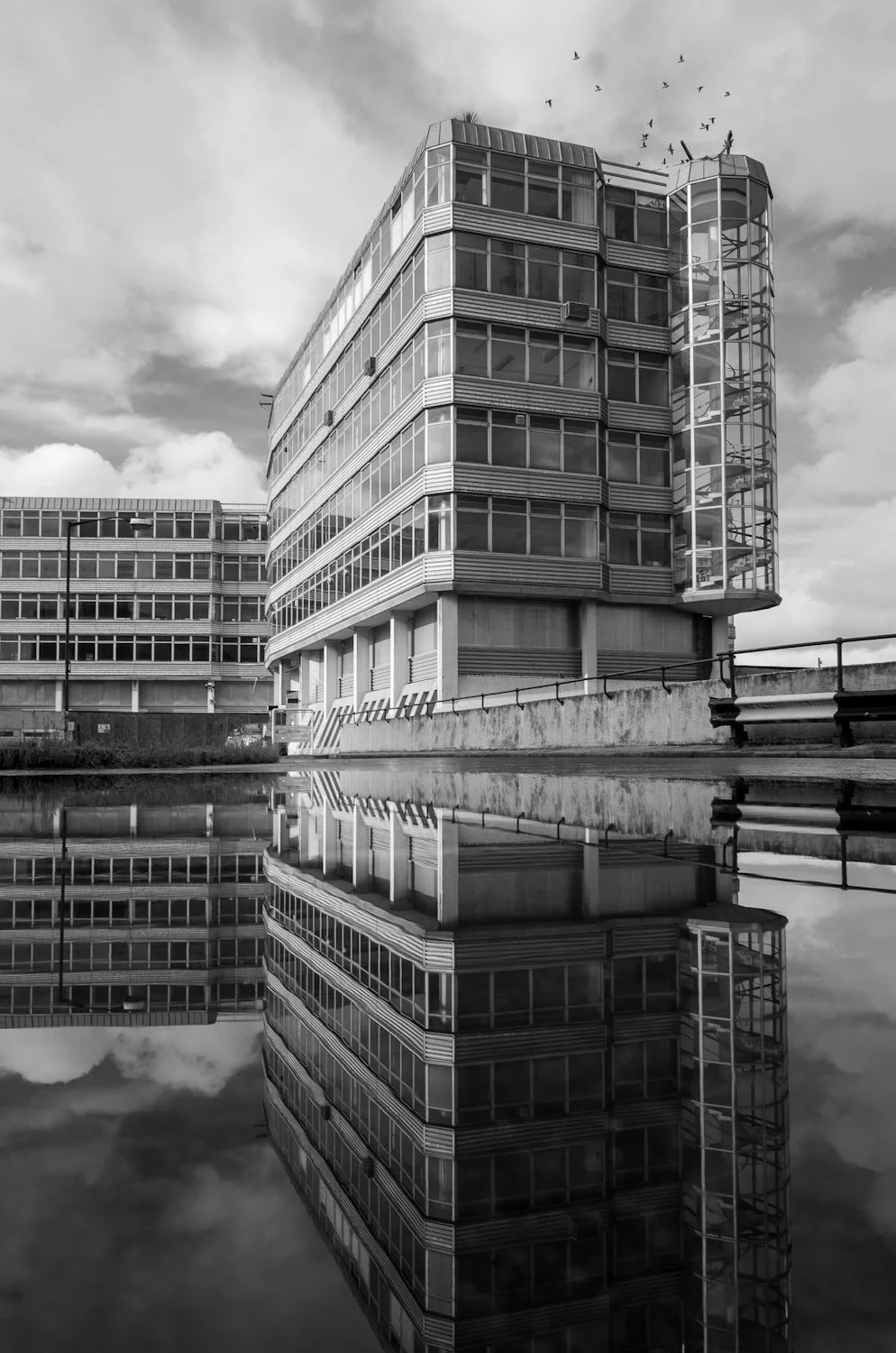 A black and white photo of a modern multi-story office building with glass windows, featuring a cylindrical spiral staircase on the corner, and a reflection of the building in a body of water in the foreground.