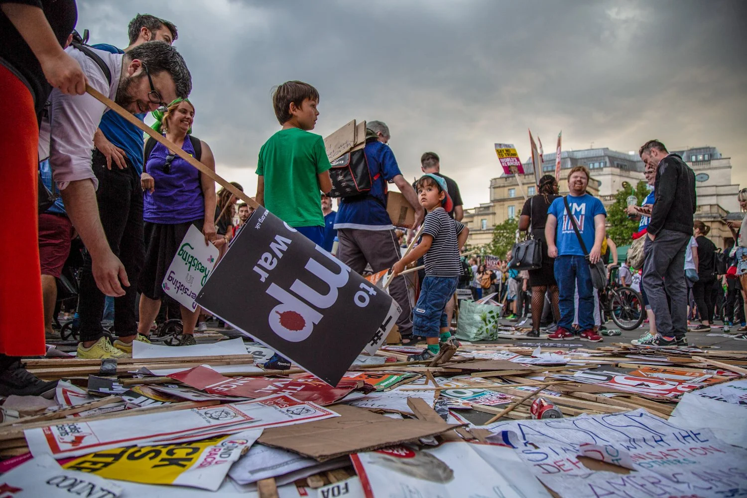 People at a protest holding signs and surrounded by scattered signs and cardboard on the ground, with a cityscape background and cloudy sky.