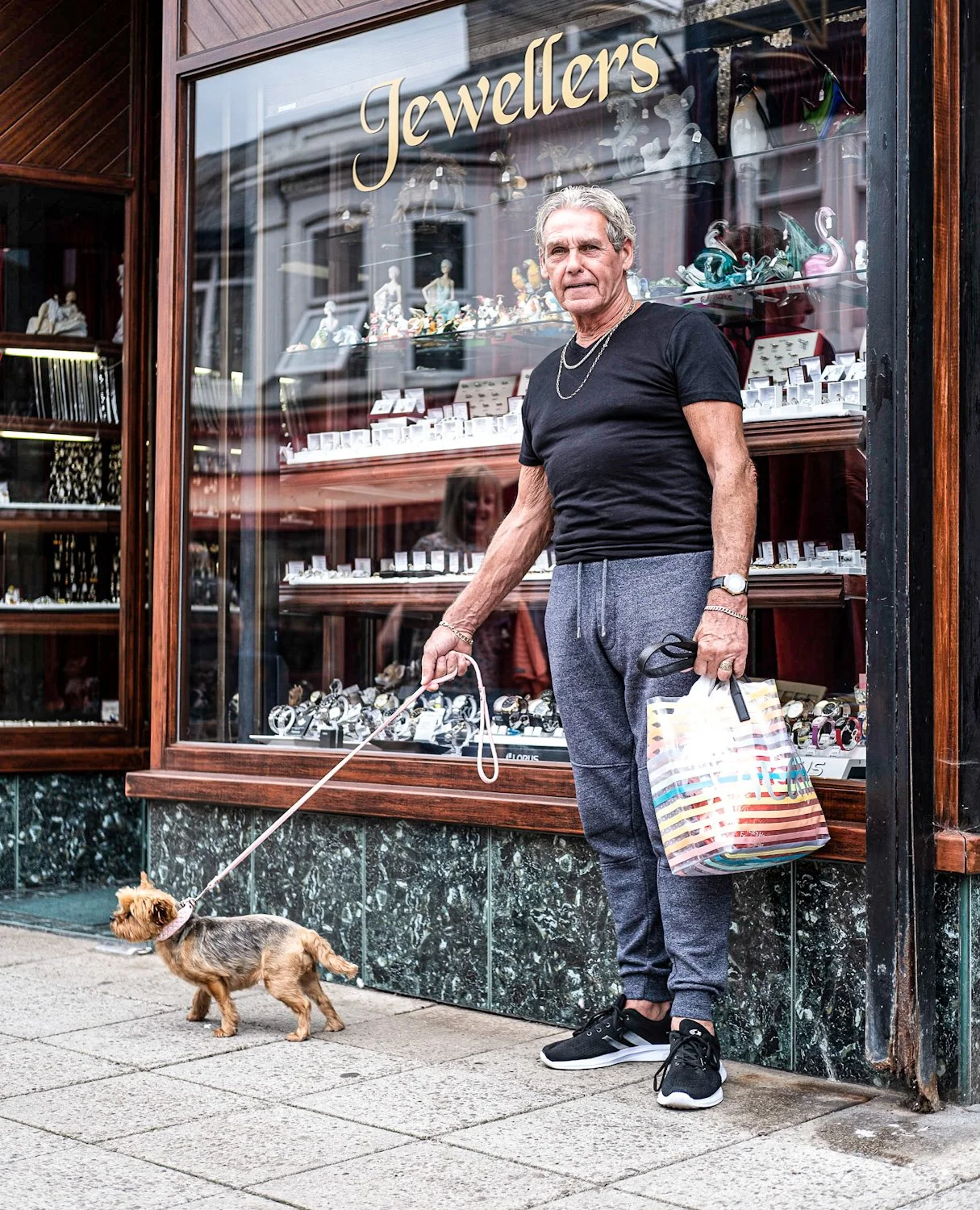 An elderly man with gray hair standing outside a jewelry store, holding a small dog on a leash in one hand and a colorful shopping bag in the other.