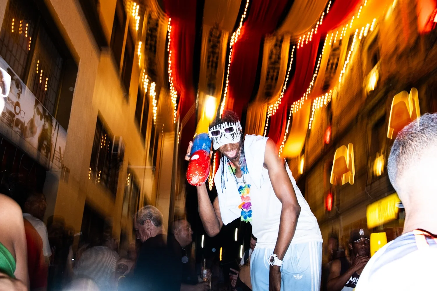Person wearing a mask, sunglasses, and colorful leis, holding a foam rocket, dancing at a lively street party or celebration at night with colorful lights and crowd.