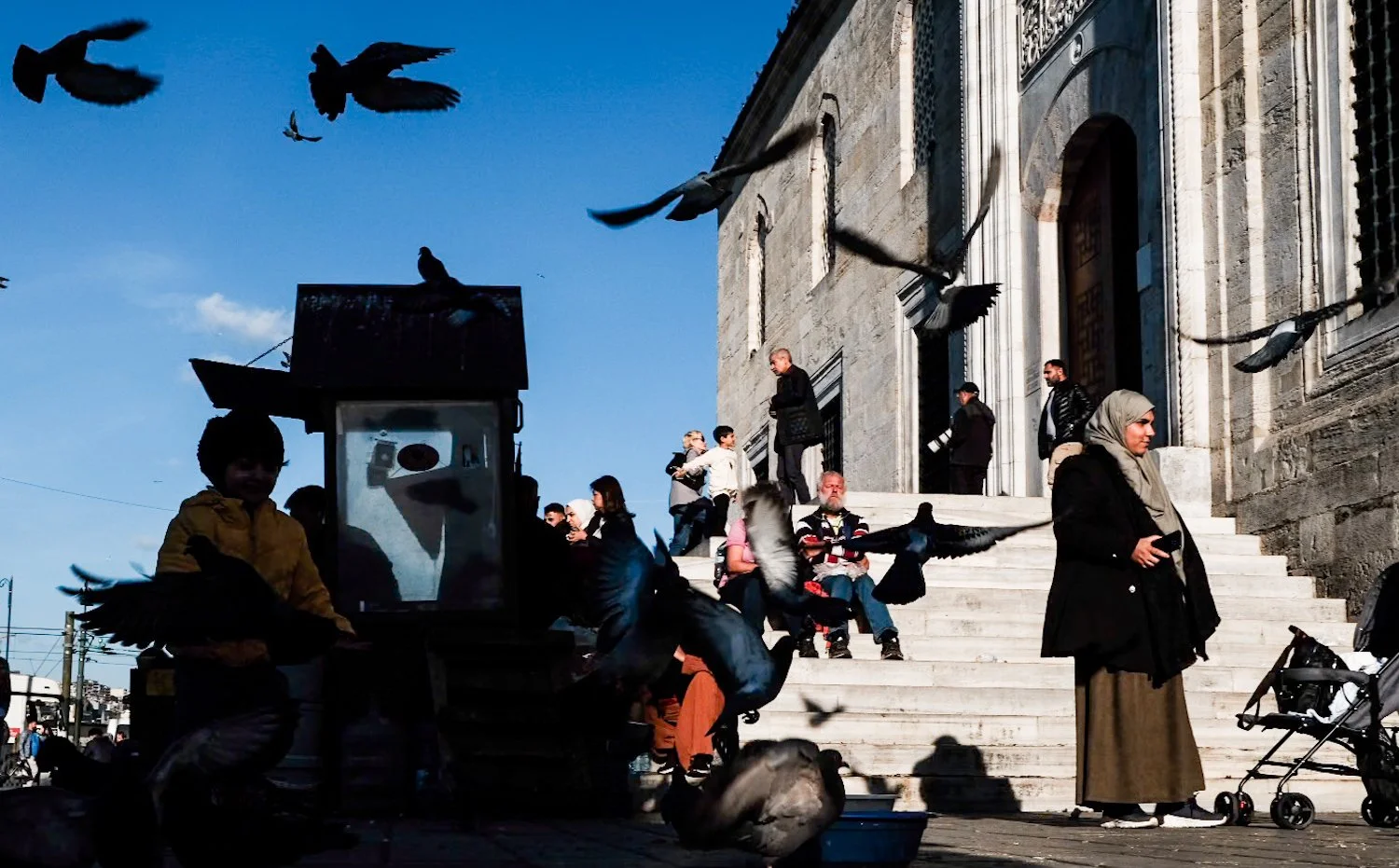 People sitting on steps outside a stone building with pigeons flying around.