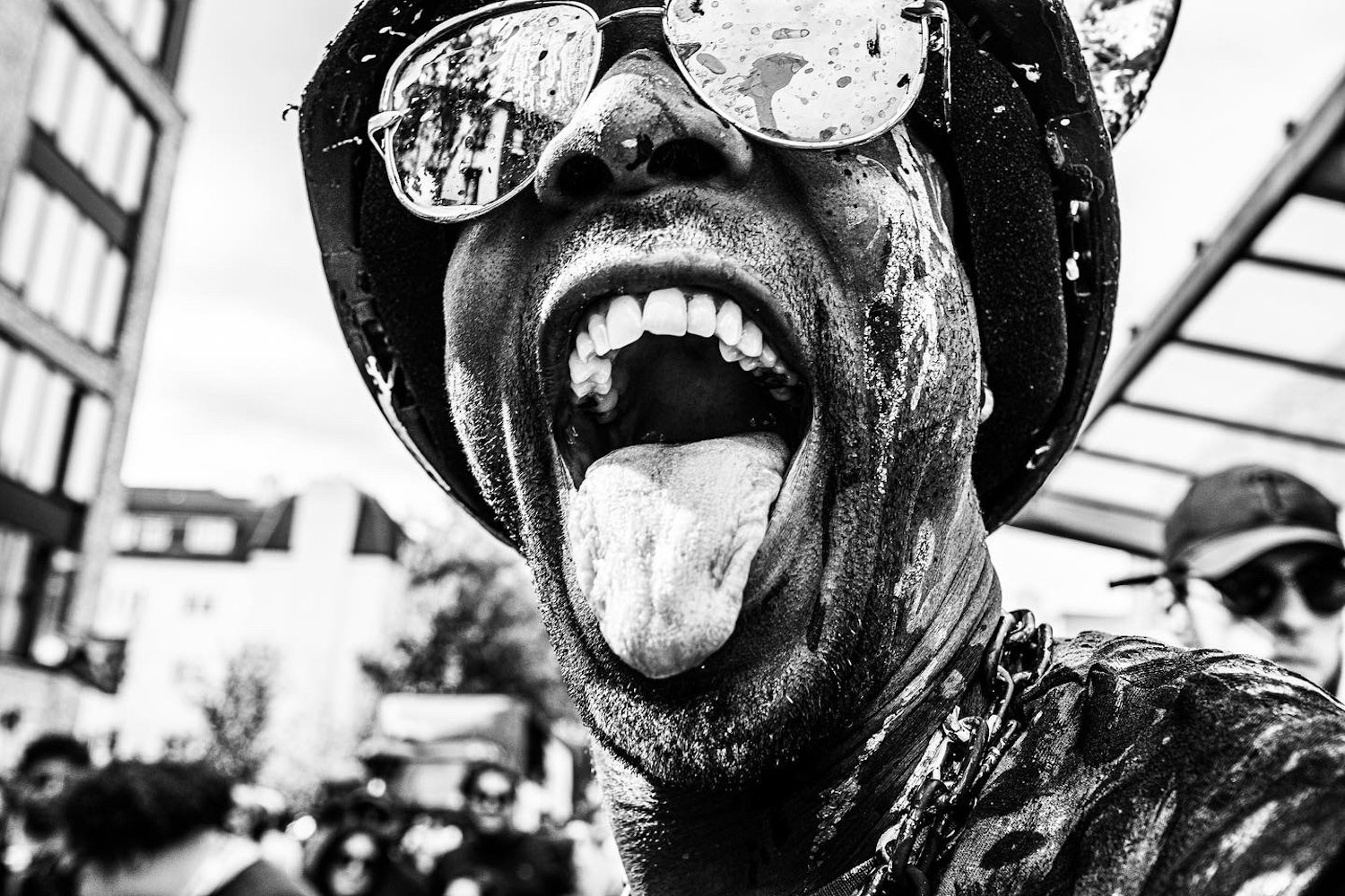 A person with sunglasses, a helmet, and a chain around their neck, sticking their tongue out, with an open mouth. The photo is black and white and appears to be taken outdoors in a crowd.