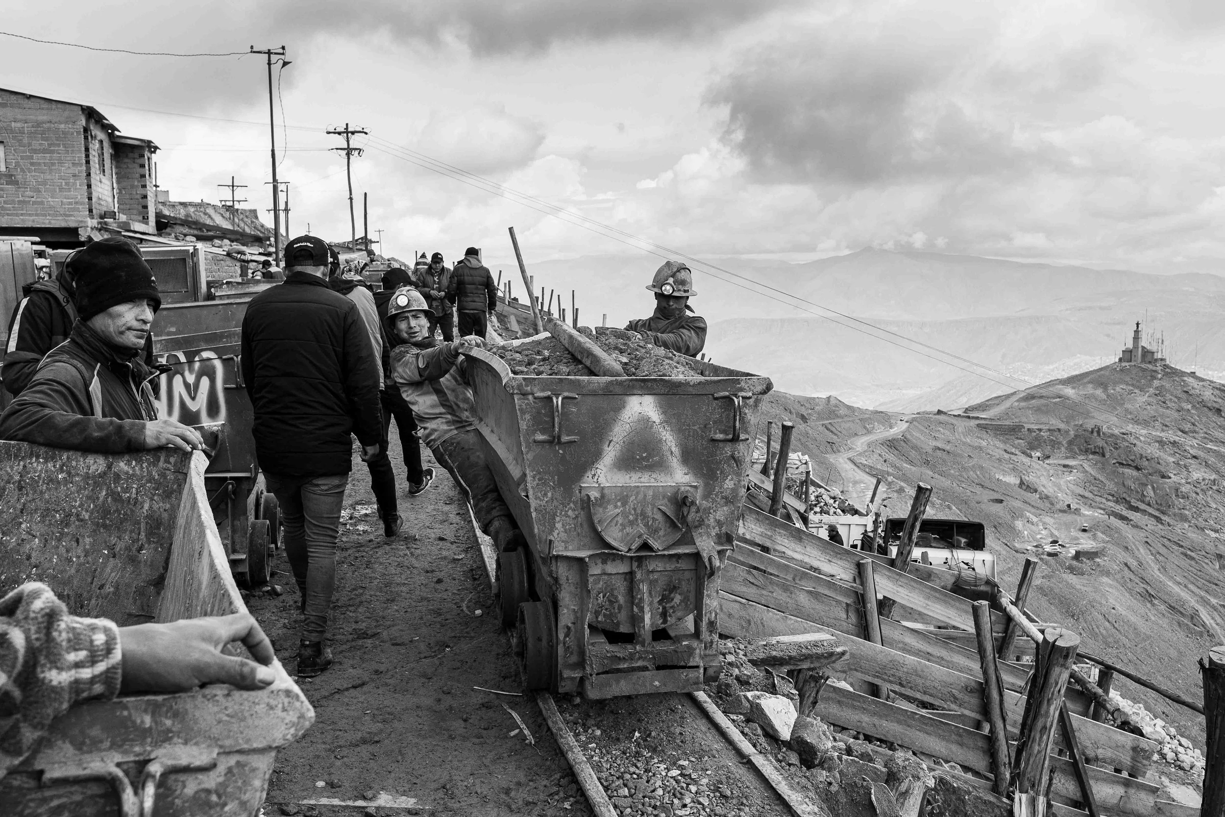 Workers in hard hats operate a bulldozer and work on a construction site on a mountainous landscape during the day, with cloudy skies overhead.