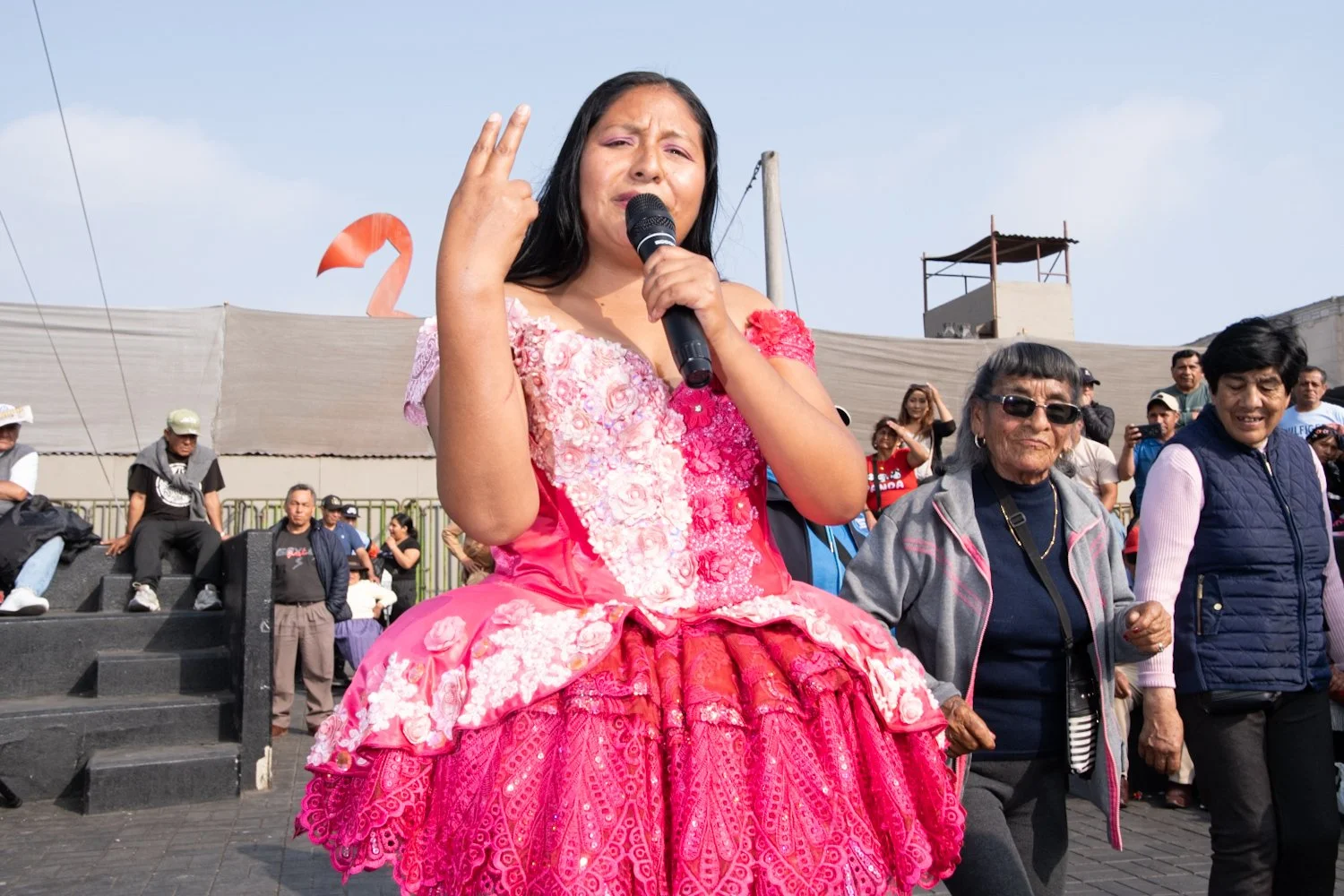 Woman wearing a bright pink, floral dress speaking into a microphone during a public event, with a diverse group of people around her.