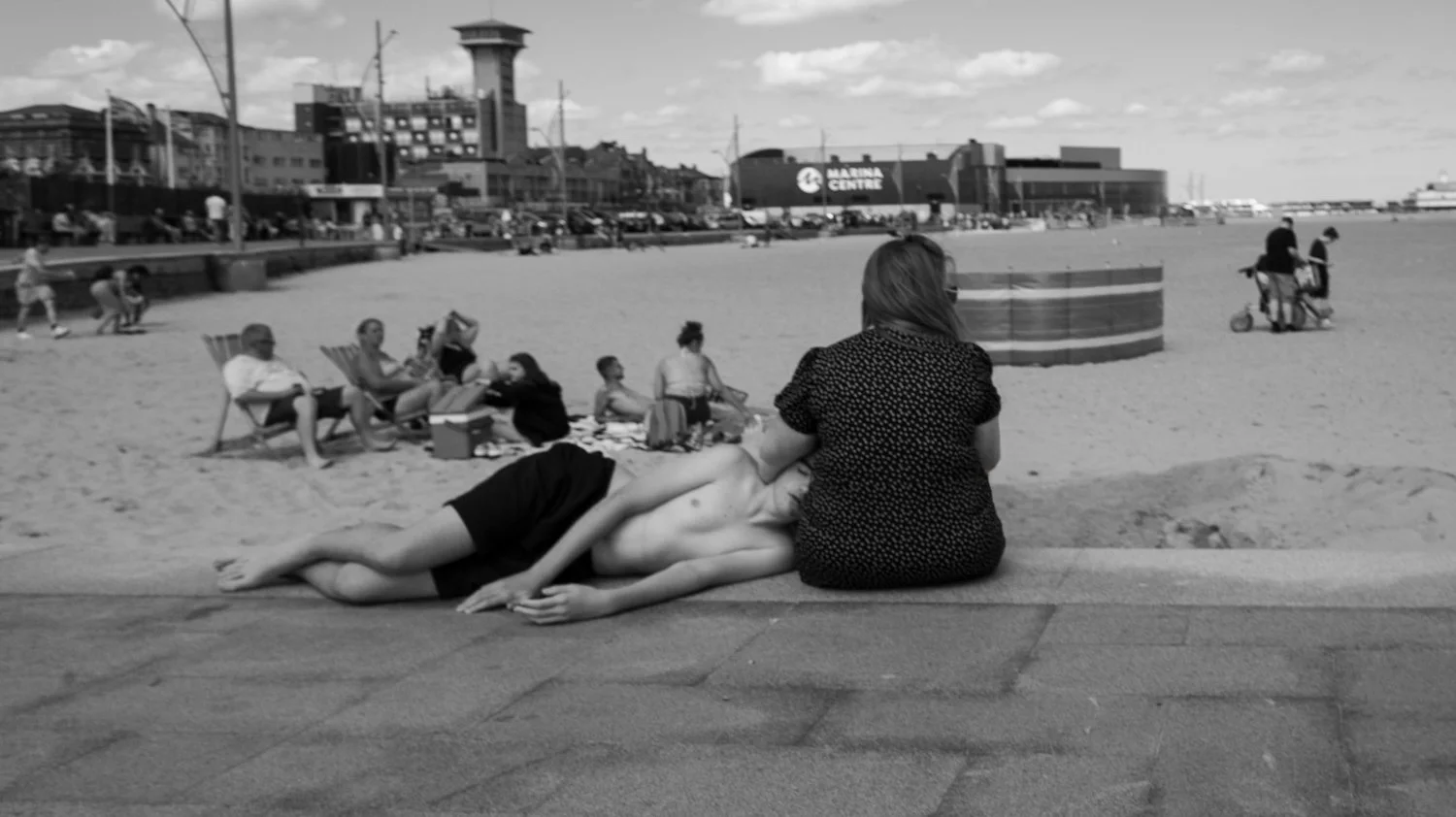 A woman peacefully sleeping on the sidewalk beside a woman sitting with her back turned, overlooking a busy beach scene with people relaxing and walking, and buildings in the background.