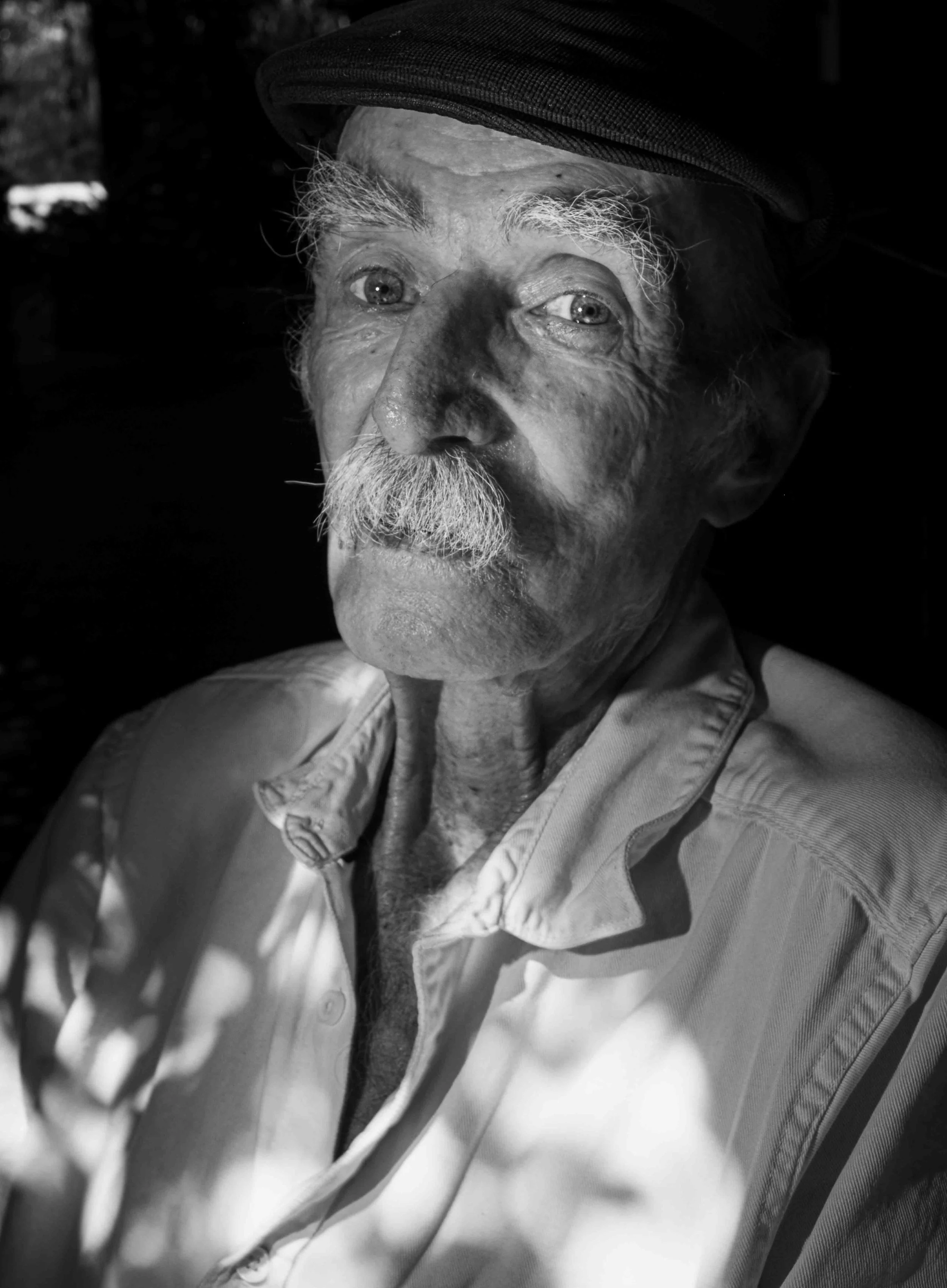 A black and white close-up portrait of an elderly man with a mustache, wearing a cap and a light-colored button-up shirt. He has light-colored eyebrows and is looking slightly to the right of the camera.