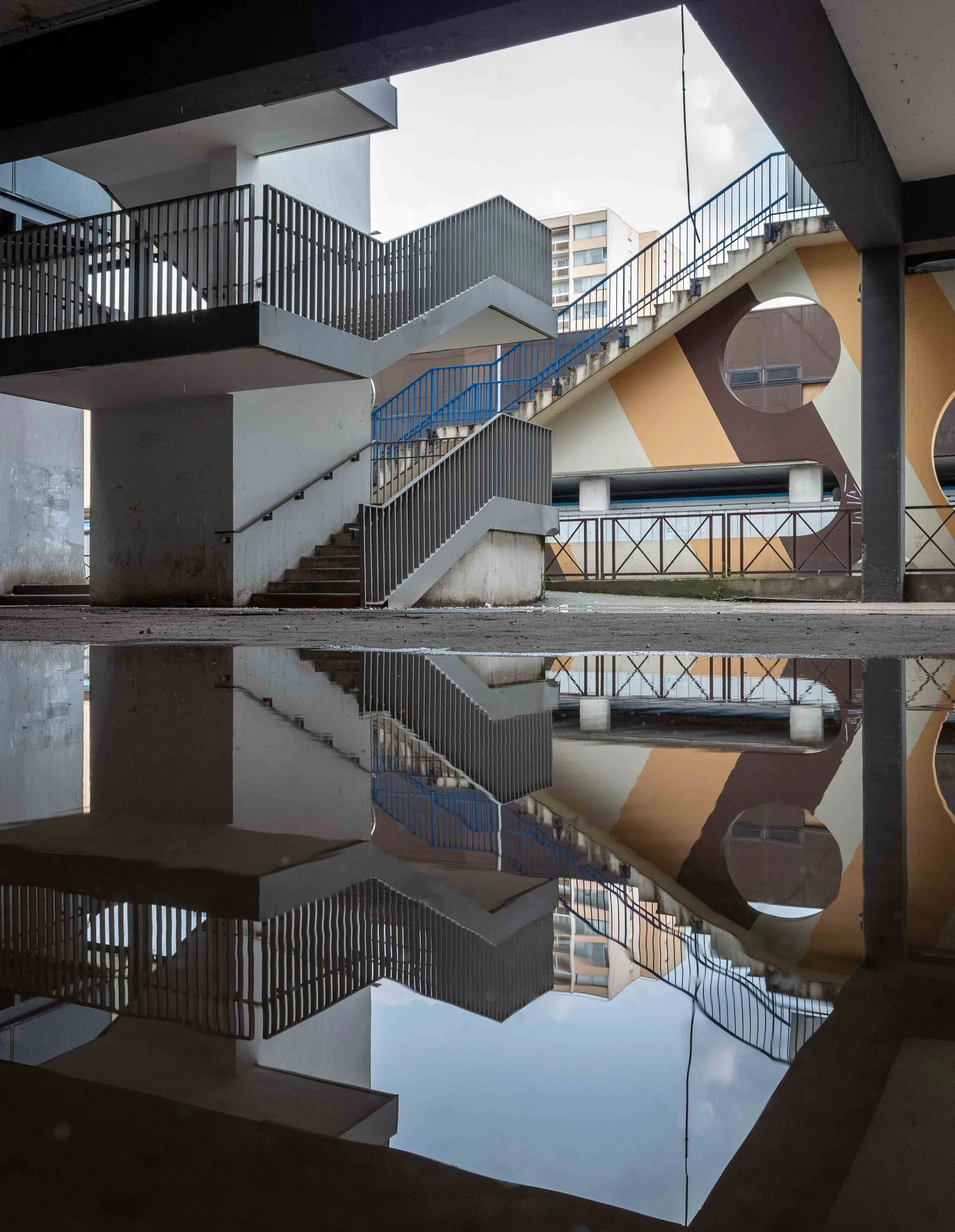 Reflection of a multi-story building with staircases and a blue railing, seen through a window with black framing during daytime, with cloudy sky overhead.
