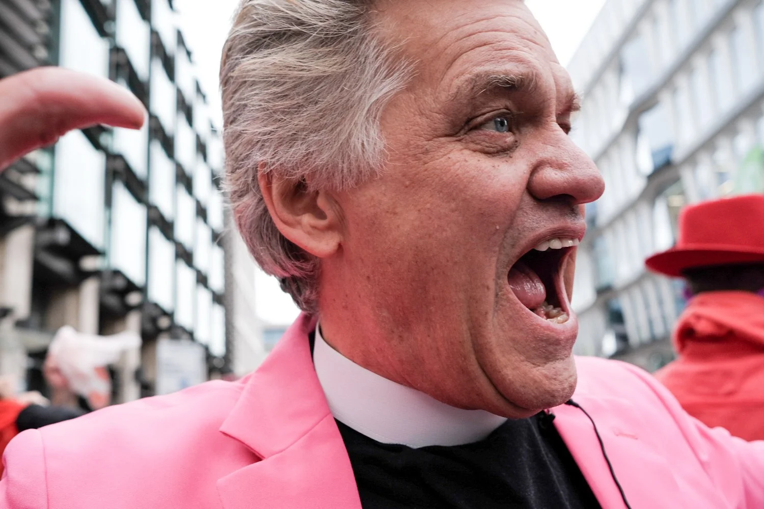 A man with gray hair, wearing a pink jacket and black shirt, appears to be yelling or cheering during an outdoor event, with a modern building in the background.