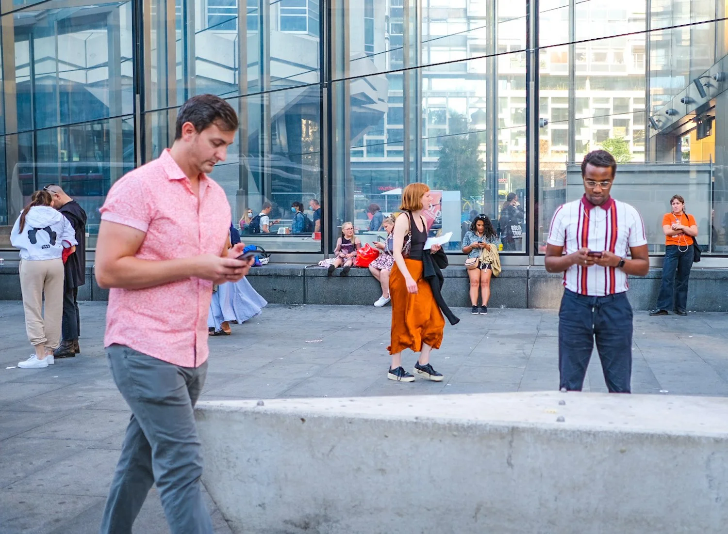 People standing and sitting outside a modern glass building, looking at their phones and reading, with some engaging in conversations.