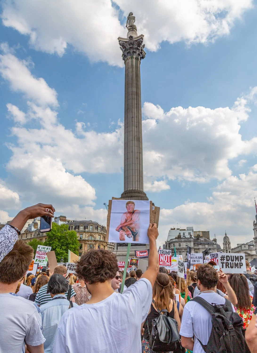 Crowd of people protesting with signs in front of Nelson's Column in Trafalgar Square, London, during daytime with partly cloudy sky.