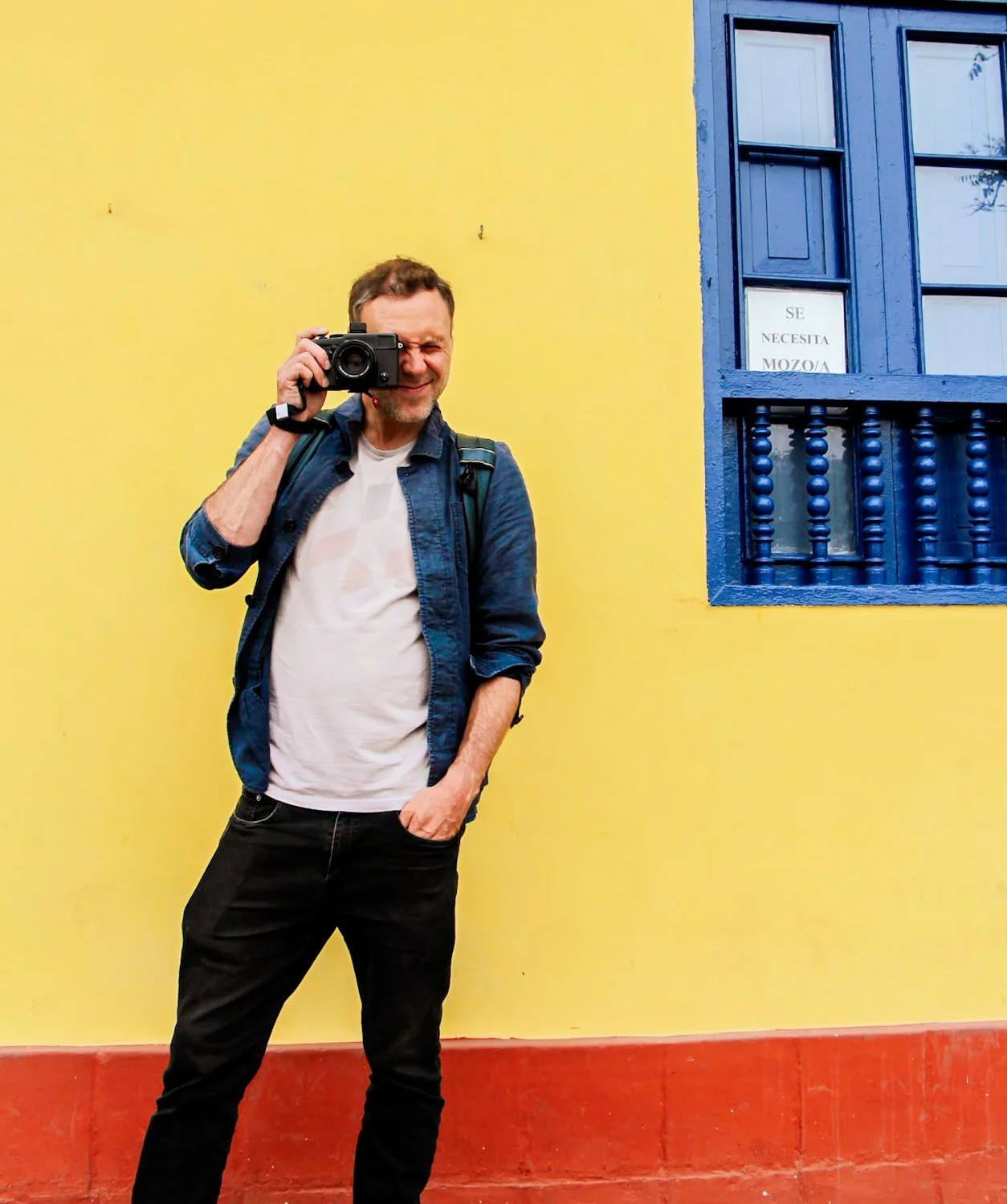 A man standing in front of a yellow wall with a blue window, holding a camera up to his eye and squinting.