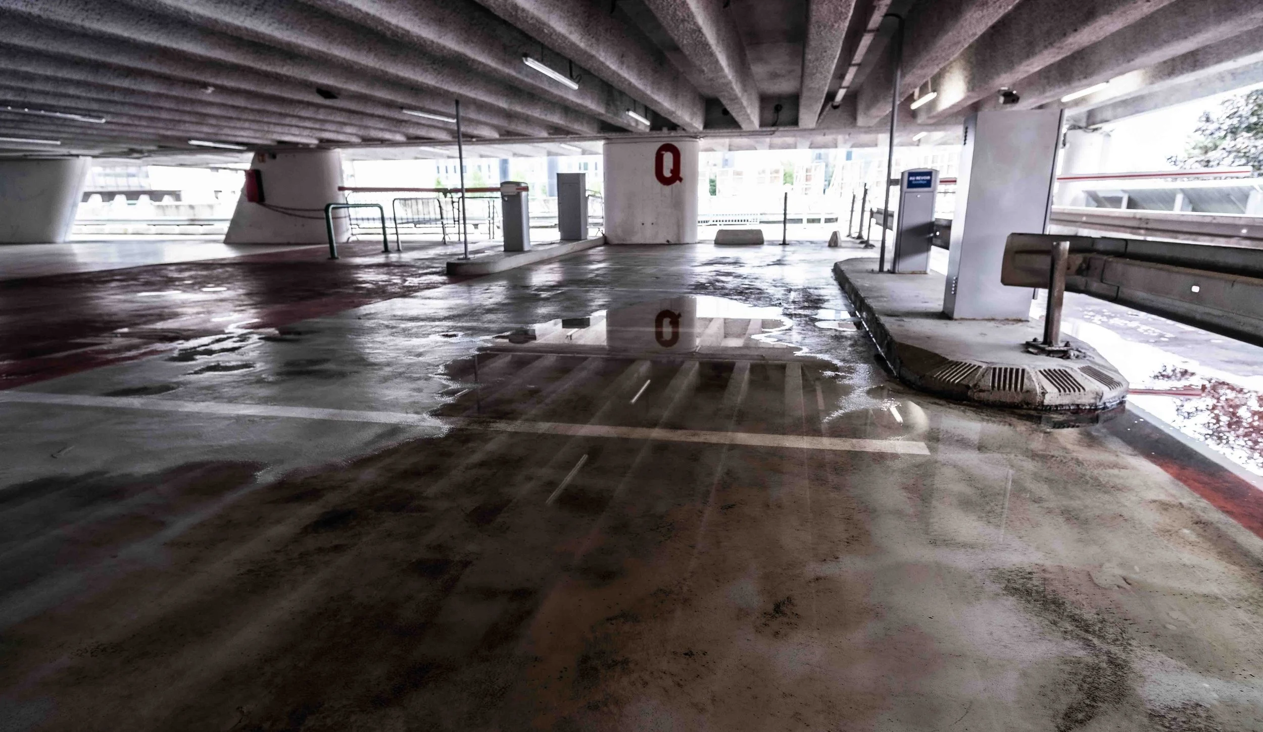 Empty parking garage with wet floor and puddles, concrete ceiling and pillars, and parked cars outside the open side.