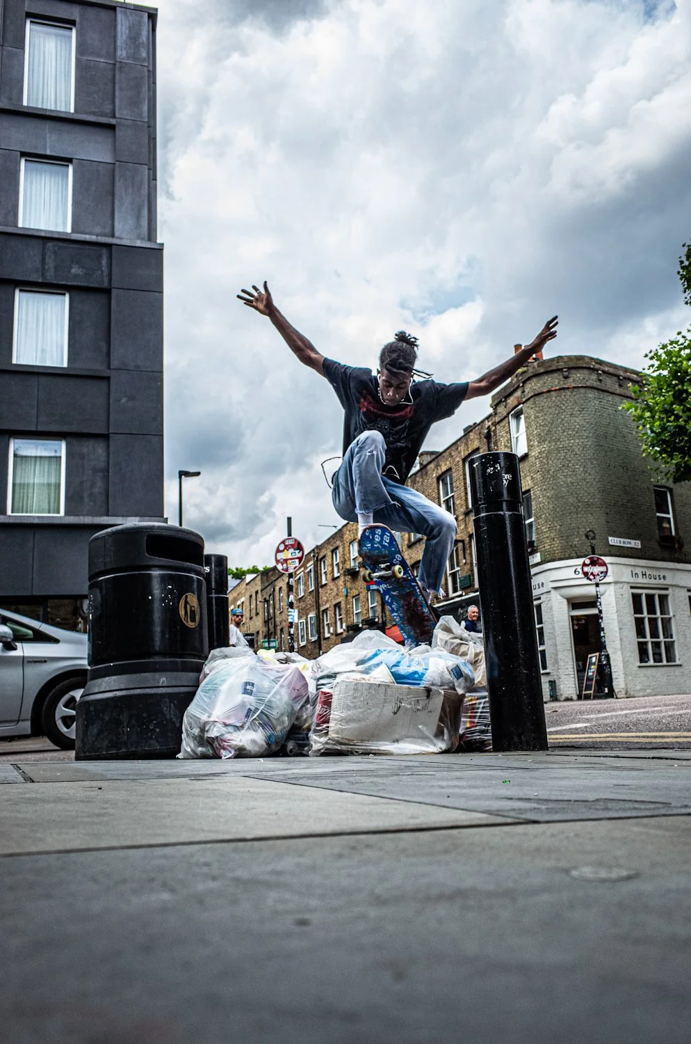 A skateboarder is performing a trick over trash bags on a city street, with buildings and cloudy sky in the background.