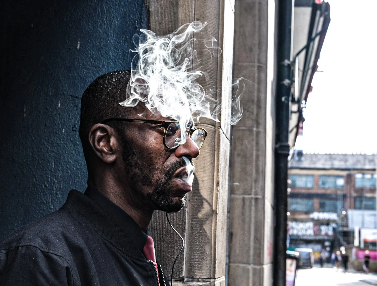 A man with glasses and a beard leaning against a wall, exhaling smoke near a city street.