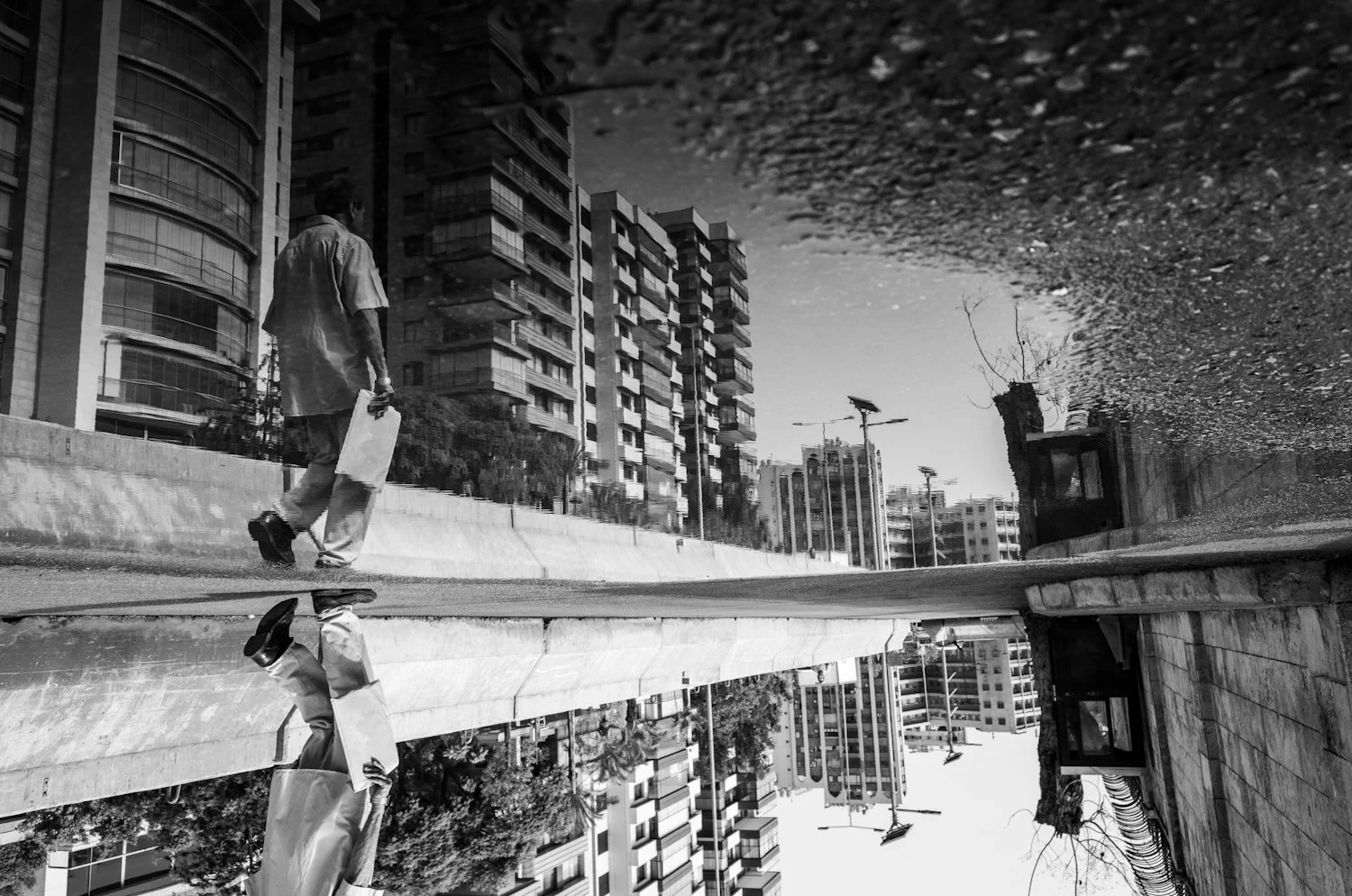 Black and white photo of a man walking on a city street carrying a paper bag, with tall apartment buildings and streetlights in the background, reflected in a puddle of water on the sidewalk.