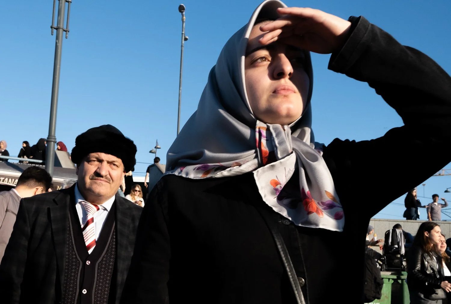 A woman wearing a headscarf shielding her eyes from the sun with her hand, standing outdoors among a crowd of people, with a man in a suit and hat behind her. The sky is clear and blue.