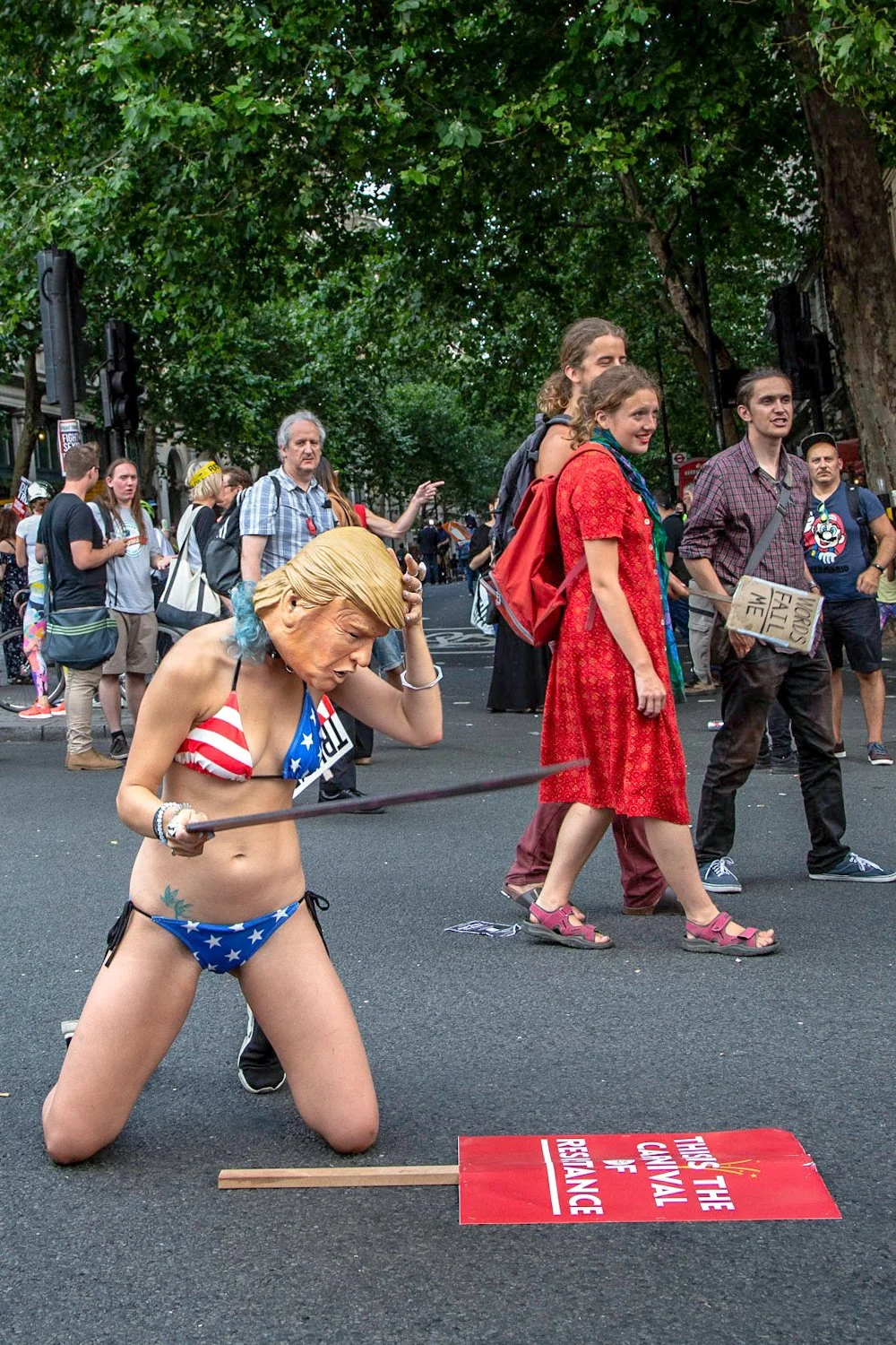 A woman dressed in a bikini with an American flag pattern is kneeling and holding a stick while participating in a protest or demonstration during a crowd event, with other people walking and engaging in conversations around her.