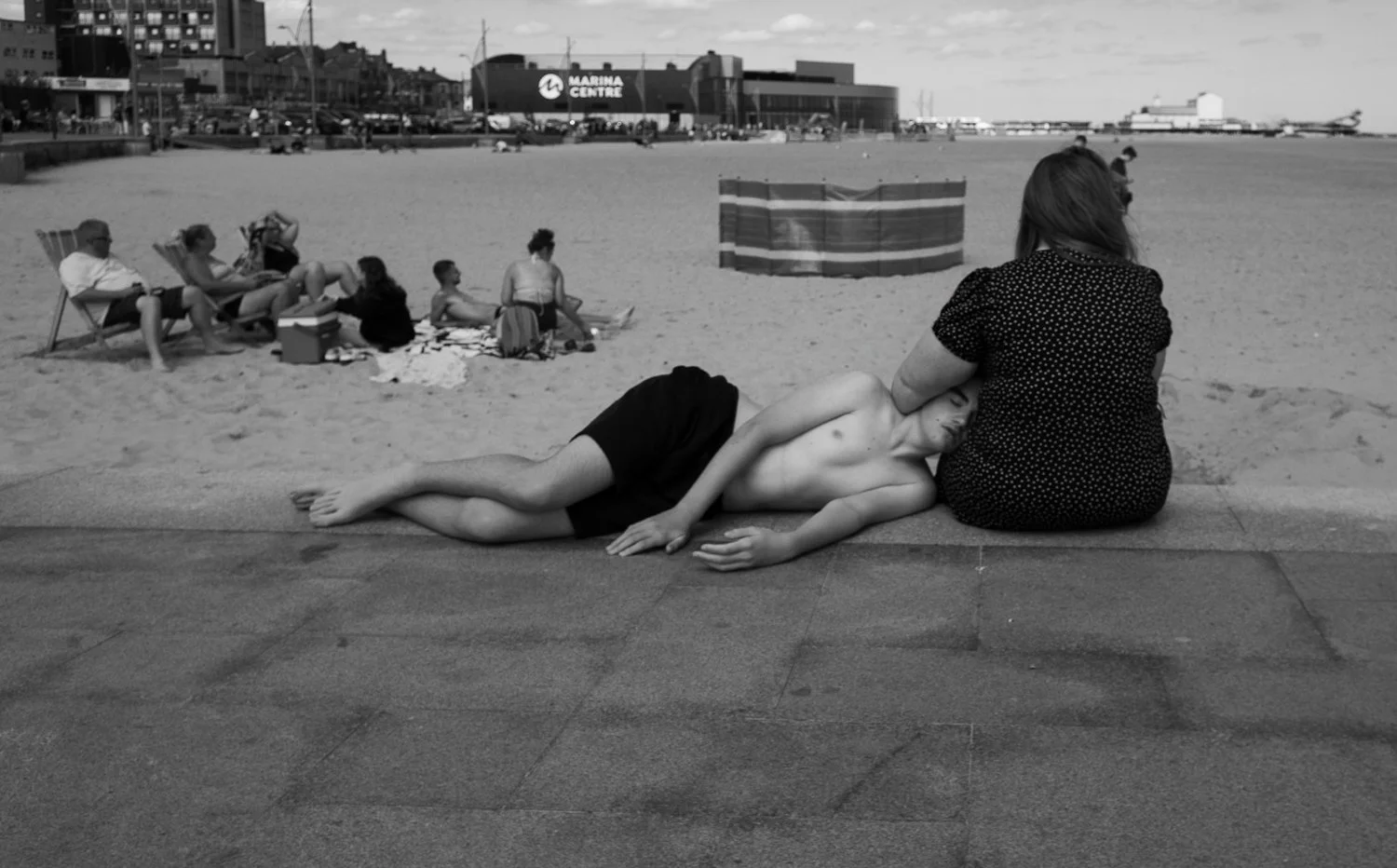 A black-and-white photo of a beach. In the foreground, a shirtless young man is lying on his side with his head resting on a woman’s lap. The woman is wearing a dotted dress and sitting on the edge of a walkway. In the background, there are several p