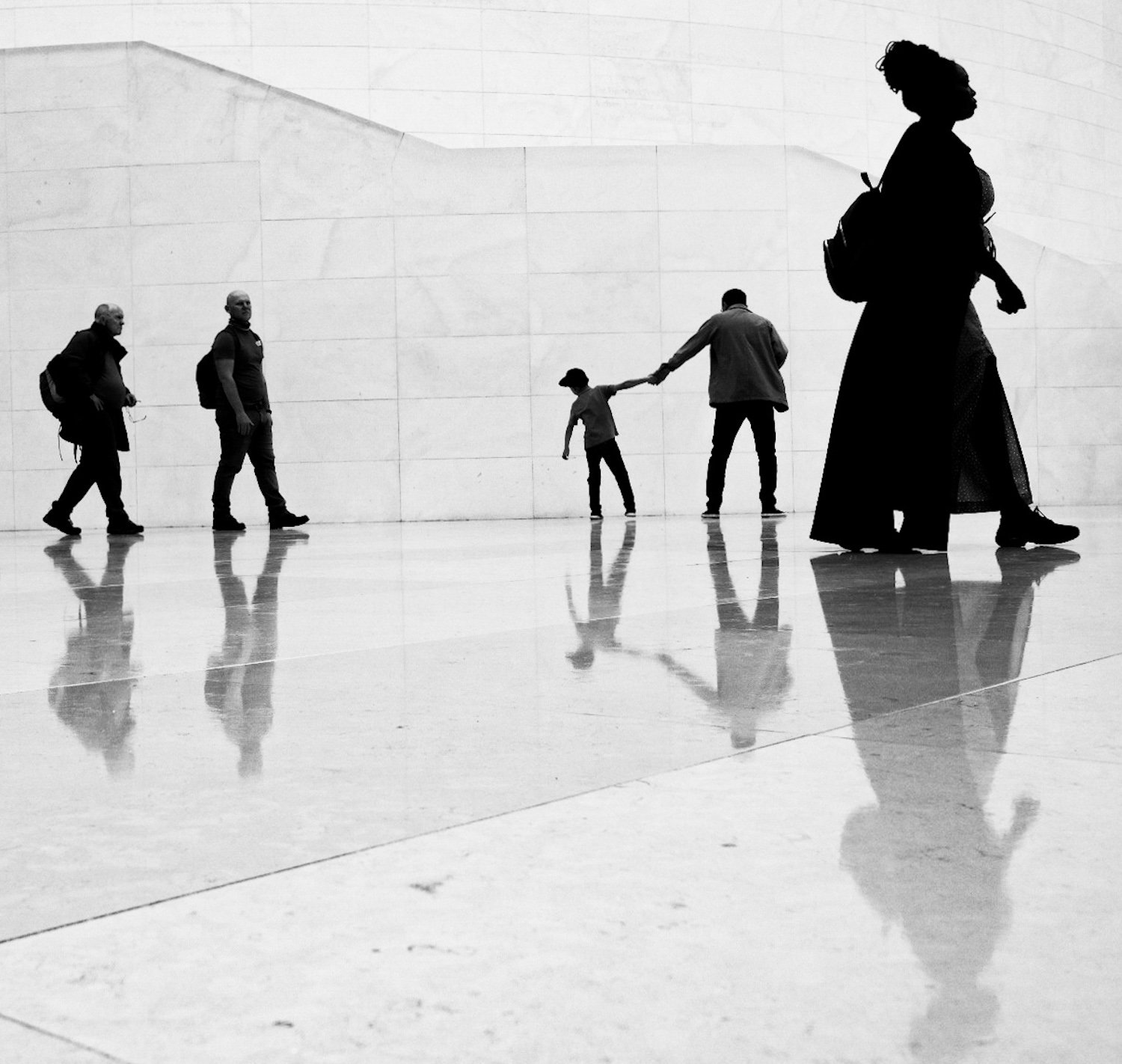 Silhouettes of six people walking in a modern indoor space with reflective floor and plain wall, including a woman with a backpack, two children holding hands, and three men in casual attire.