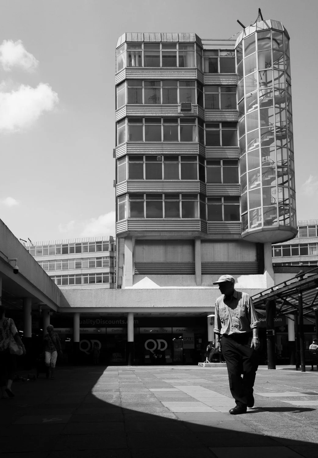 A black and white photo of a modern multi-story building with a glass spiral staircase on the right side, and a man walking in the foreground.