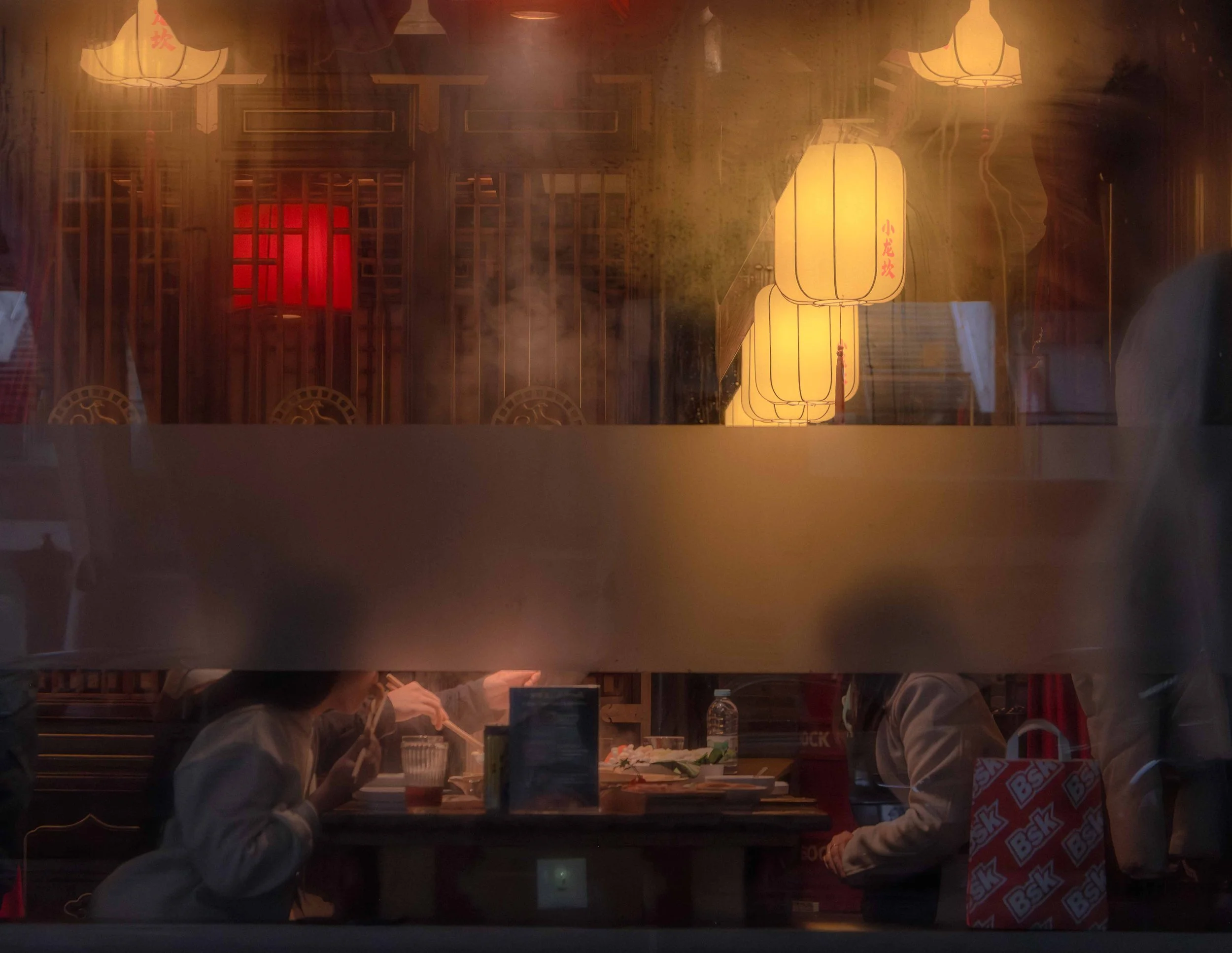 People dining at a restaurant with Japanese decor, wooden lattice walls, red window accents, paper lanterns with Japanese writing, and steam rising from the kitchen.