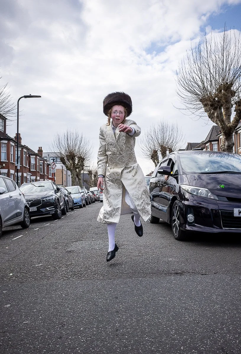 A person in a fancy gold patterned coat, white tights, black shoes, and a large hat, jumping in the middle of a street lined with parked cars and leafless trees, with a cloudy sky above.