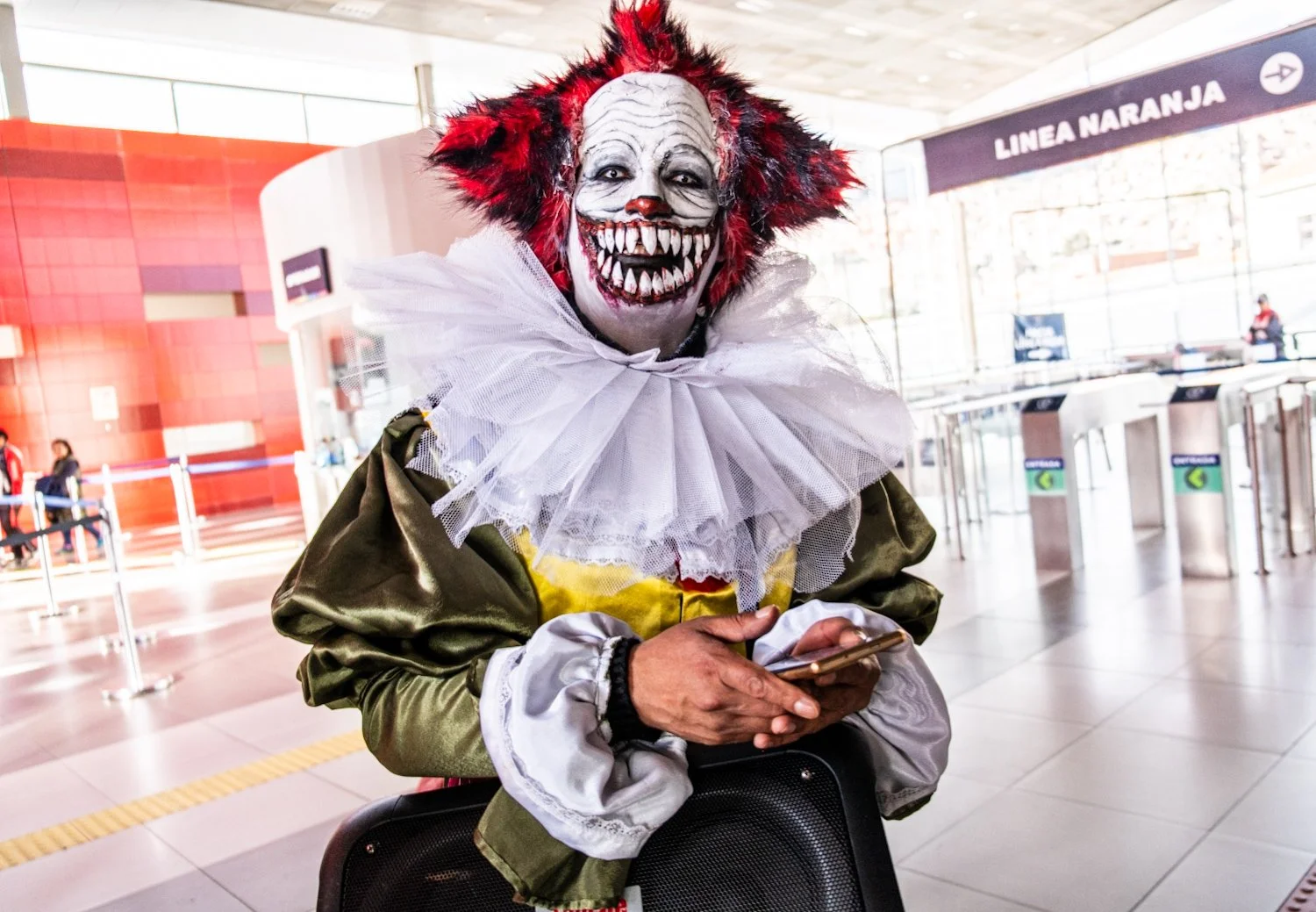 Person dressed as Pennywise clown from 'It' movie with clown makeup, red hair, white ruffled collar, using a smartphone near an airport check-in area.
