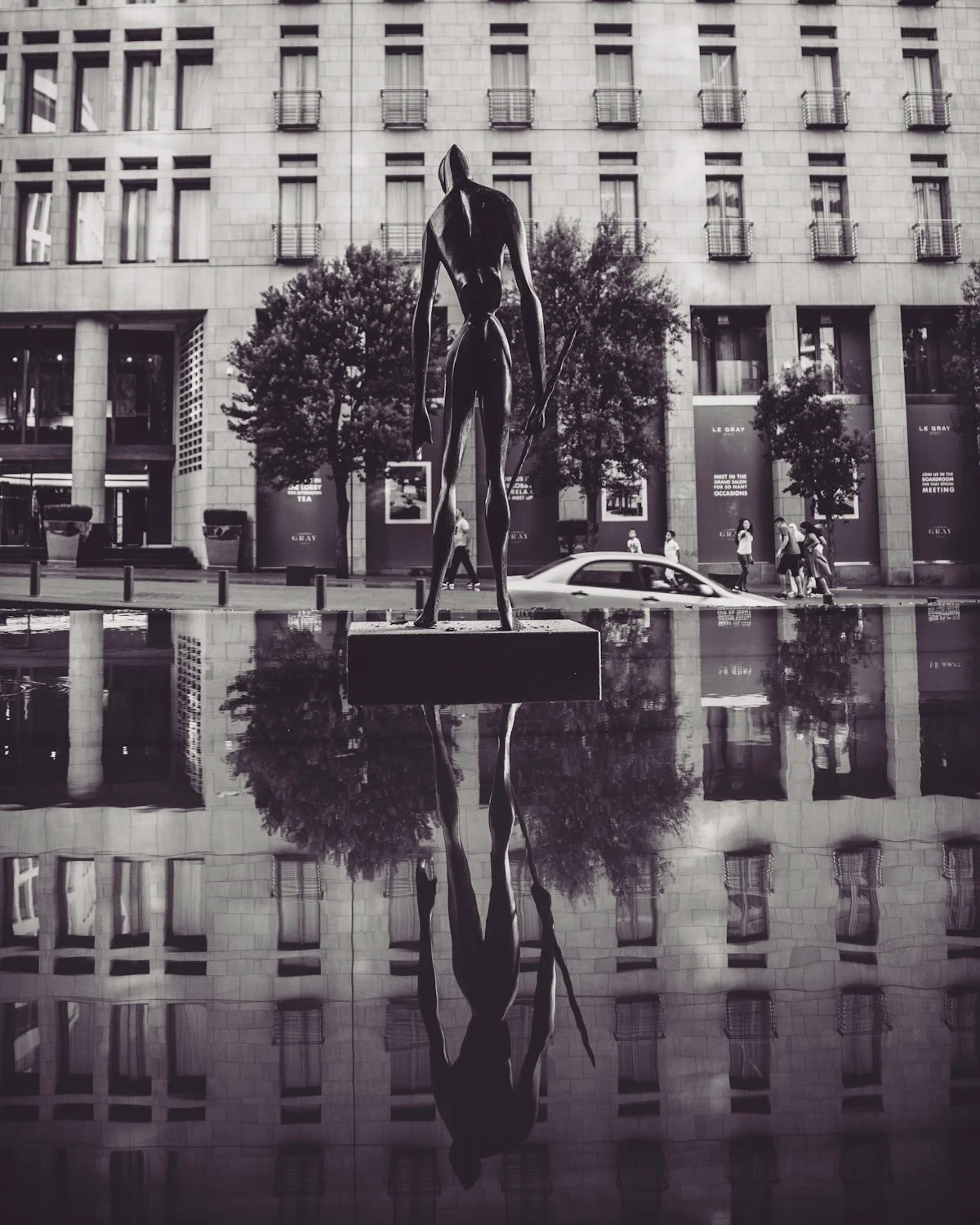 A black-and-white photo of a modern city scene featuring a sculpture of a tall, slender figure holding a spear, reflected in a water feature in the foreground. Behind, a building with multiple windows and balconies, and people walking on the sidewalk