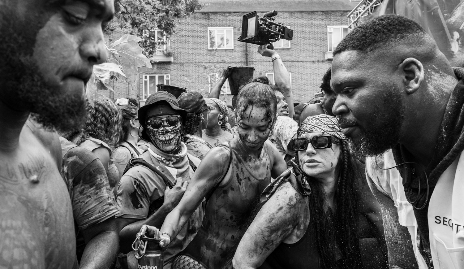 Black and white photo of a group of people at a protest or rally, with some covered in mud or dirt, engaging with a security officer or organizer, in an outdoor urban setting with trees and brick buildings.