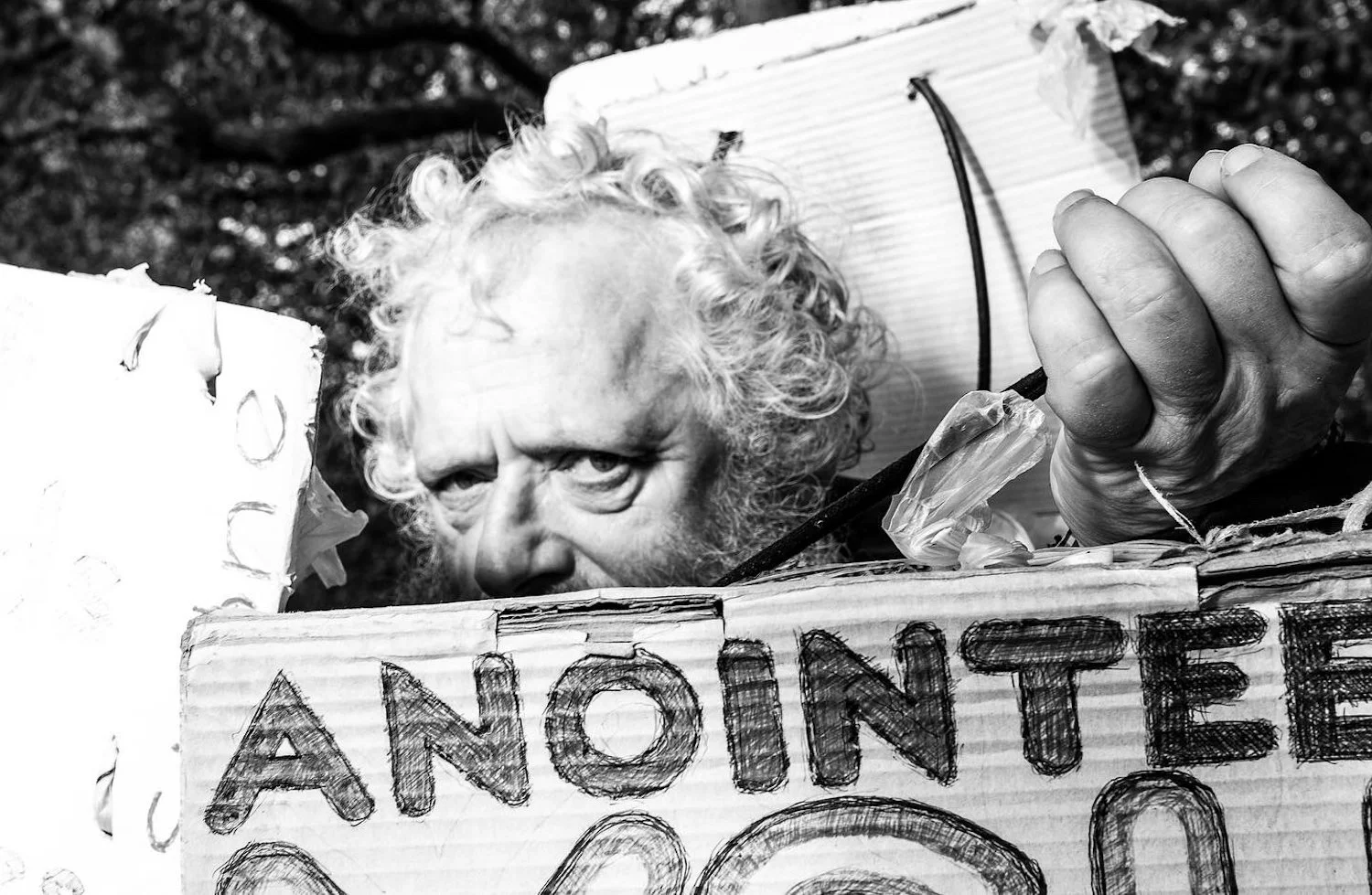 A person with curly hair looking over a protest sign that says "AN INTE" in black marker on cardboard, with a background of trees.