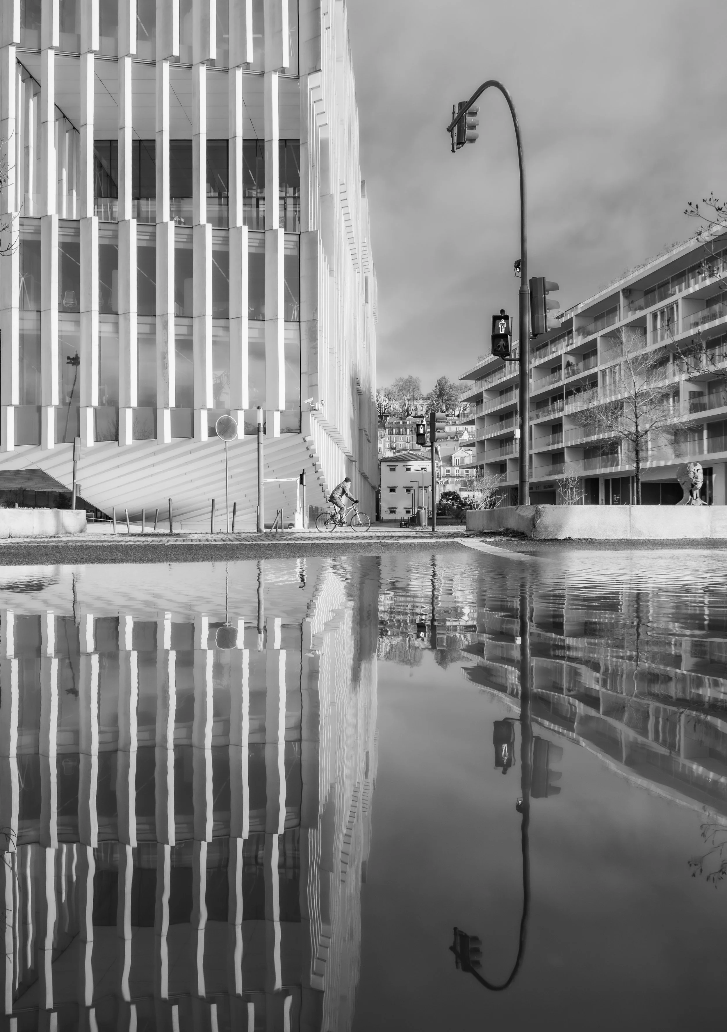 A black and white photo of modern office buildings reflecting on a puddle of water at an intersection with traffic lights, a person riding a bike, and a fountain statue in the background.
